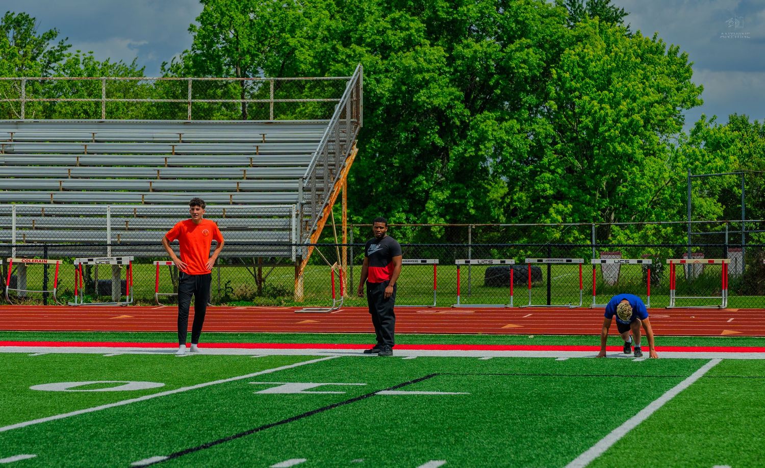 Coaches and athletes training on a red track and green field with bleachers and trees in background.