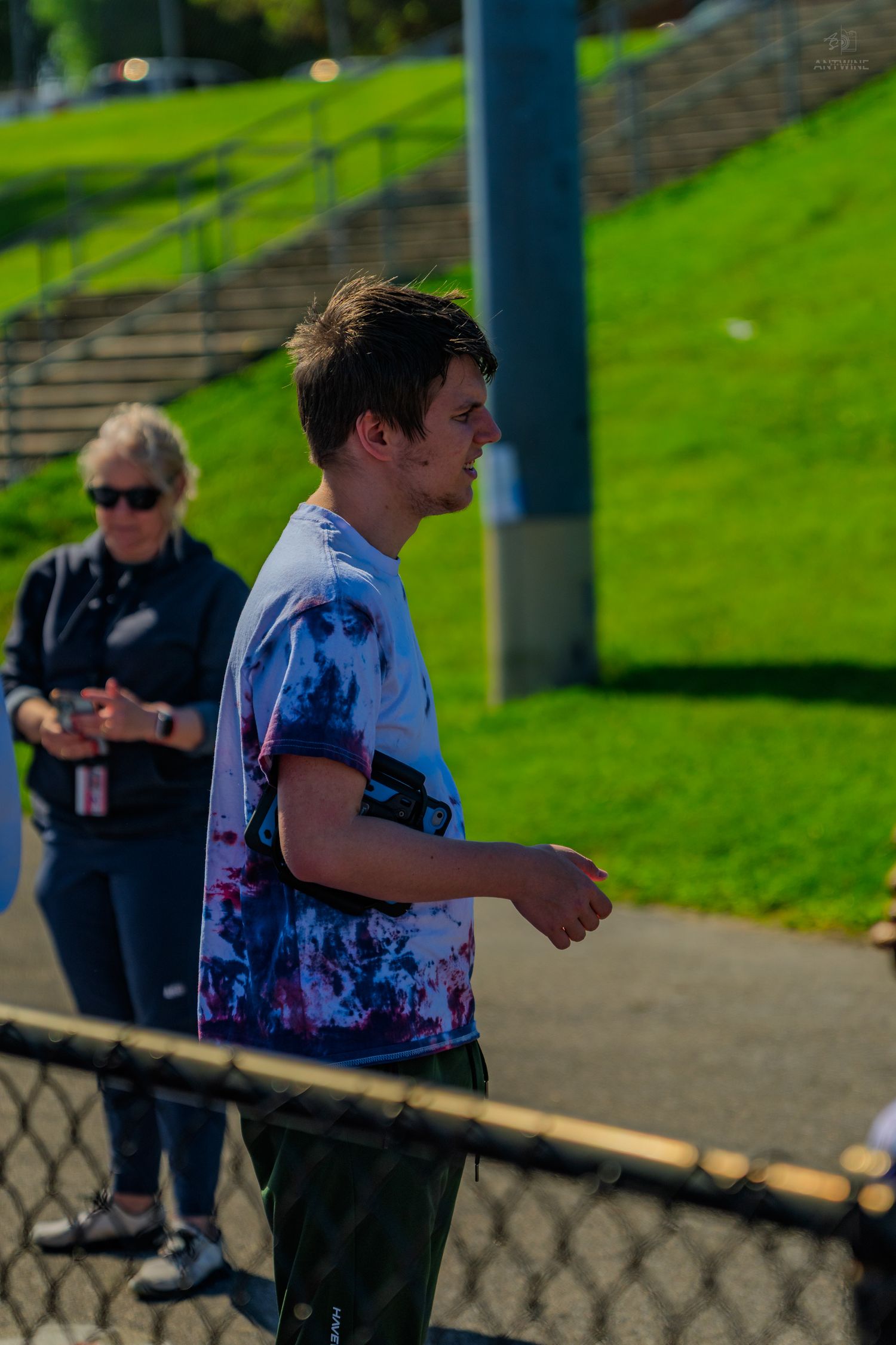 Young man in tie-dye shirt with phone armband walks near a fence at an outdoor sports venue on a sunny day.