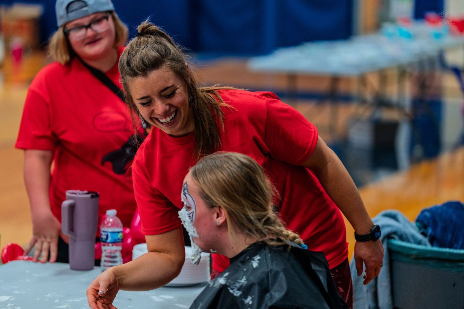Two women in red shirts laugh together at a table during a fun indoor event or activity.
