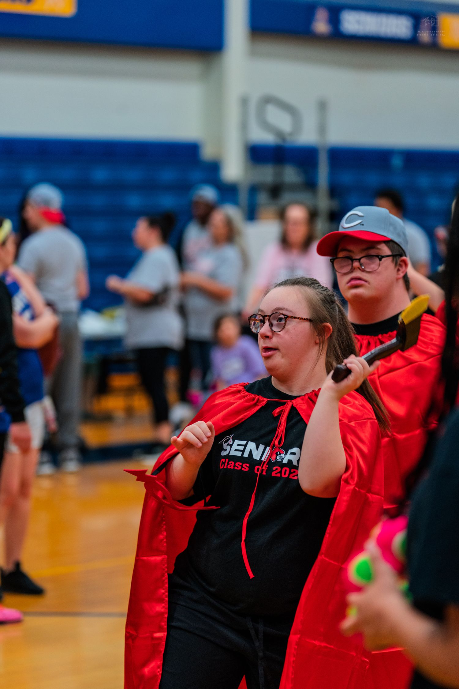 High school seniors wearing red capes and black Senior Class 2023 shirts participate in a school gymnasium celebration.