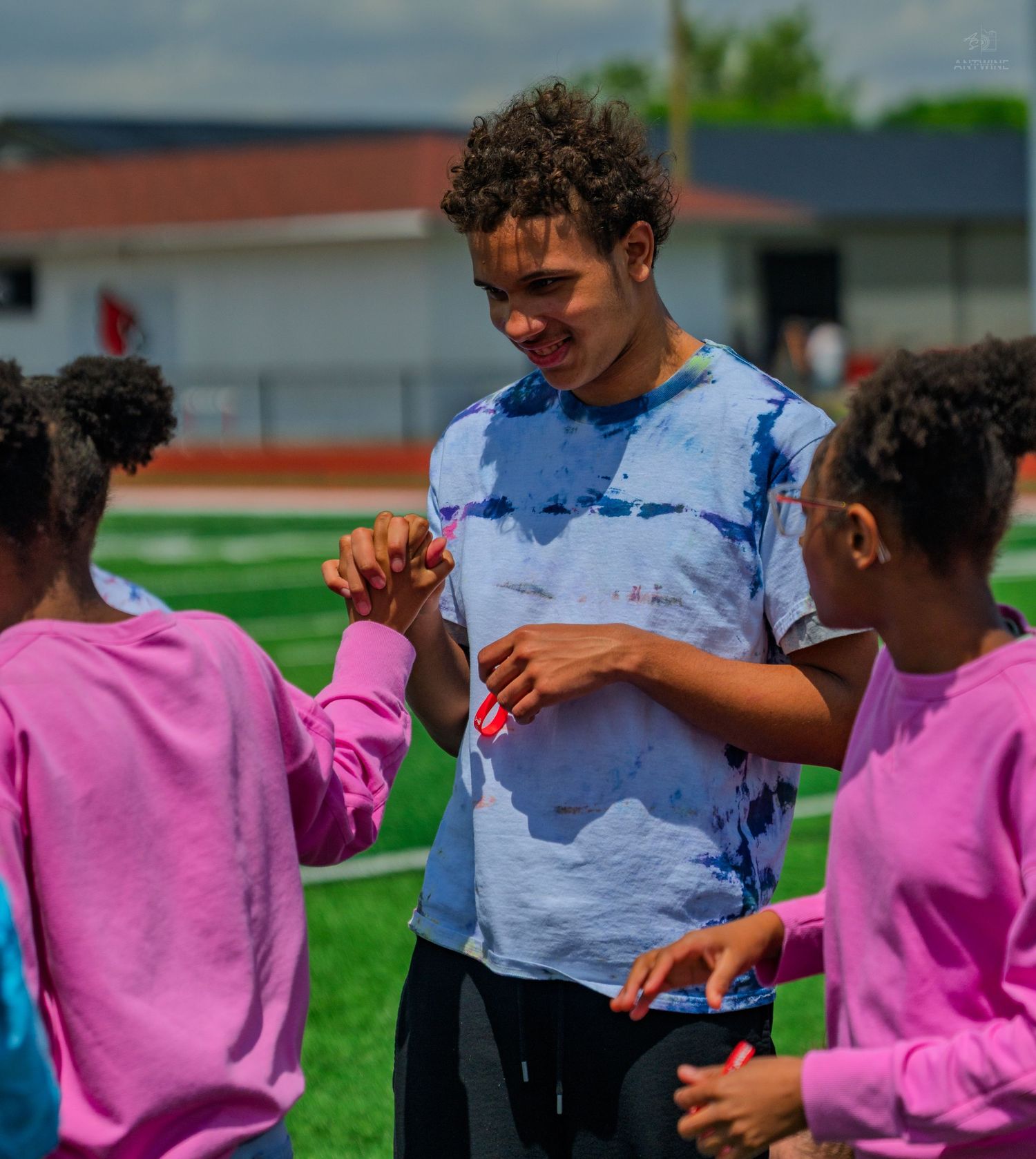 Teen boy in tie-dye shirt fist-bumping young girls in pink sweatshirts on a green sports field.