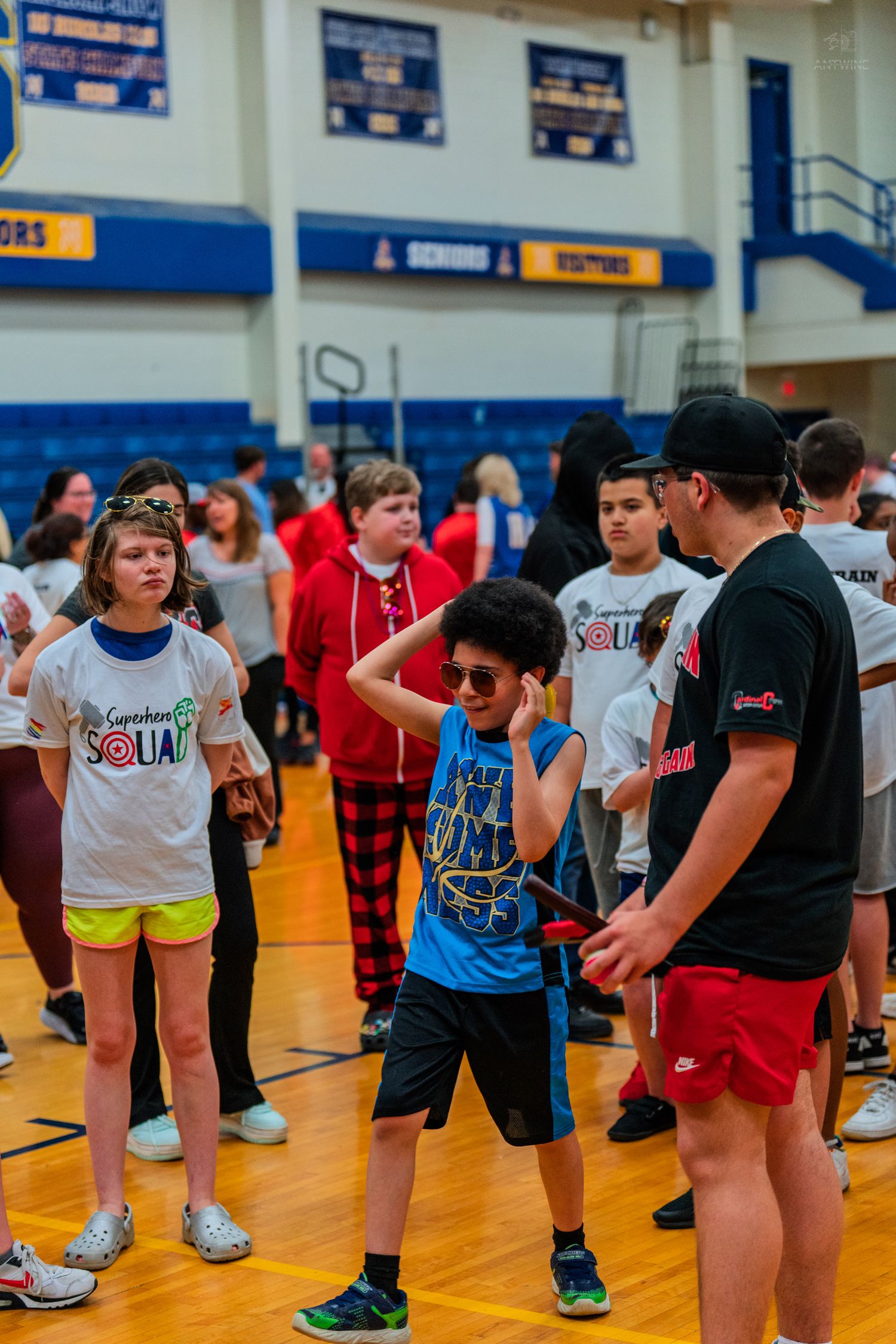 Kids participate in a school gym event, with a boy in a blue shirt and afro wig posing confidently.