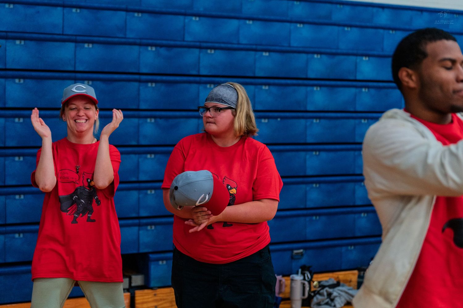 Two women in red t-shirts clap and cheer enthusiastically in a gymnasium during a school event.