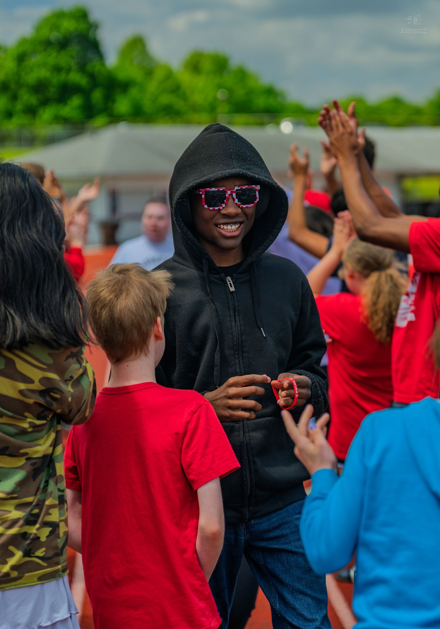 Smiling teen in black hoodie and pixel sunglasses enjoys outdoor event surrounded by kids in red shirts.