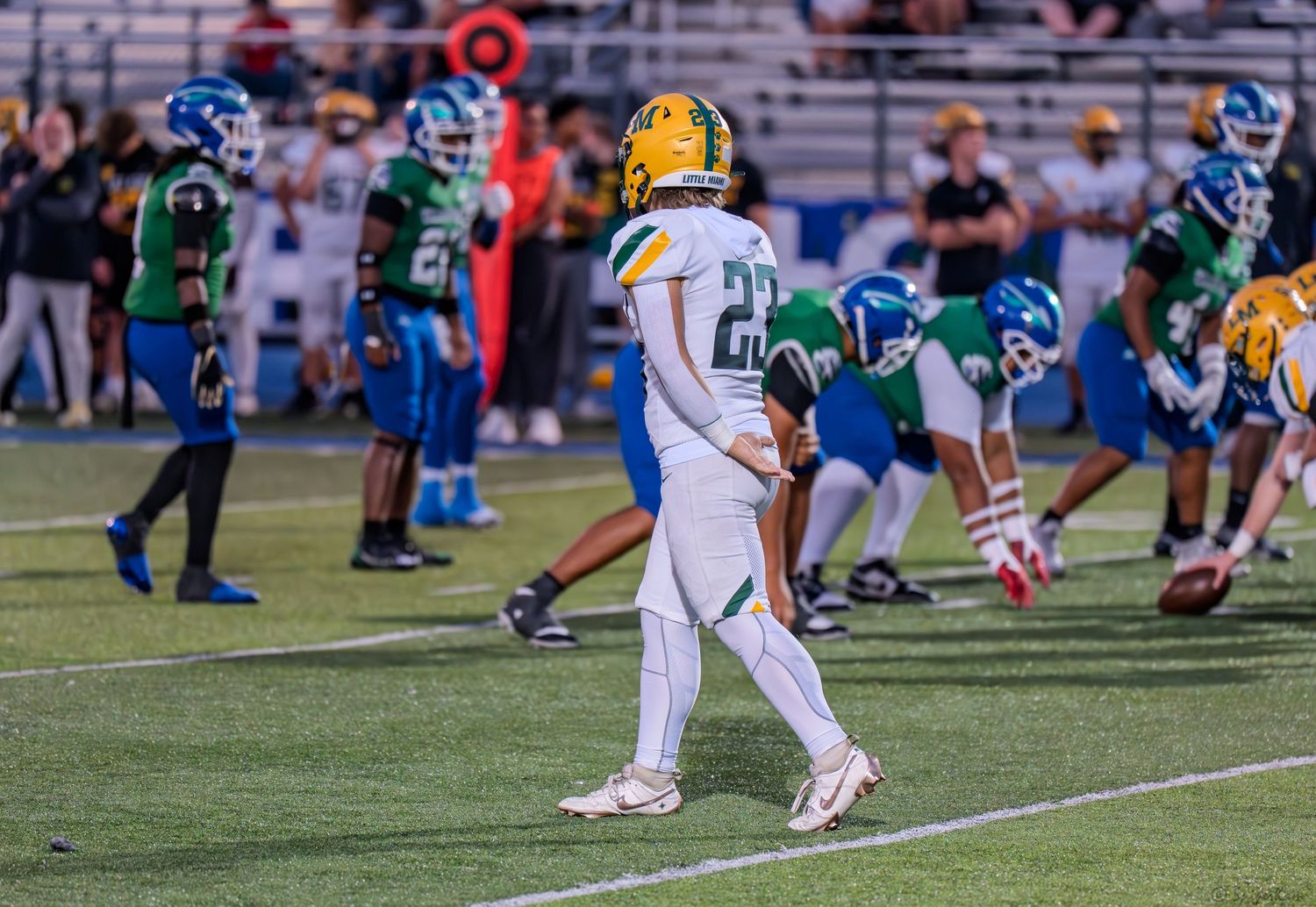 High school football player number 22 in white and gold uniform walks on field during night game against teal team.