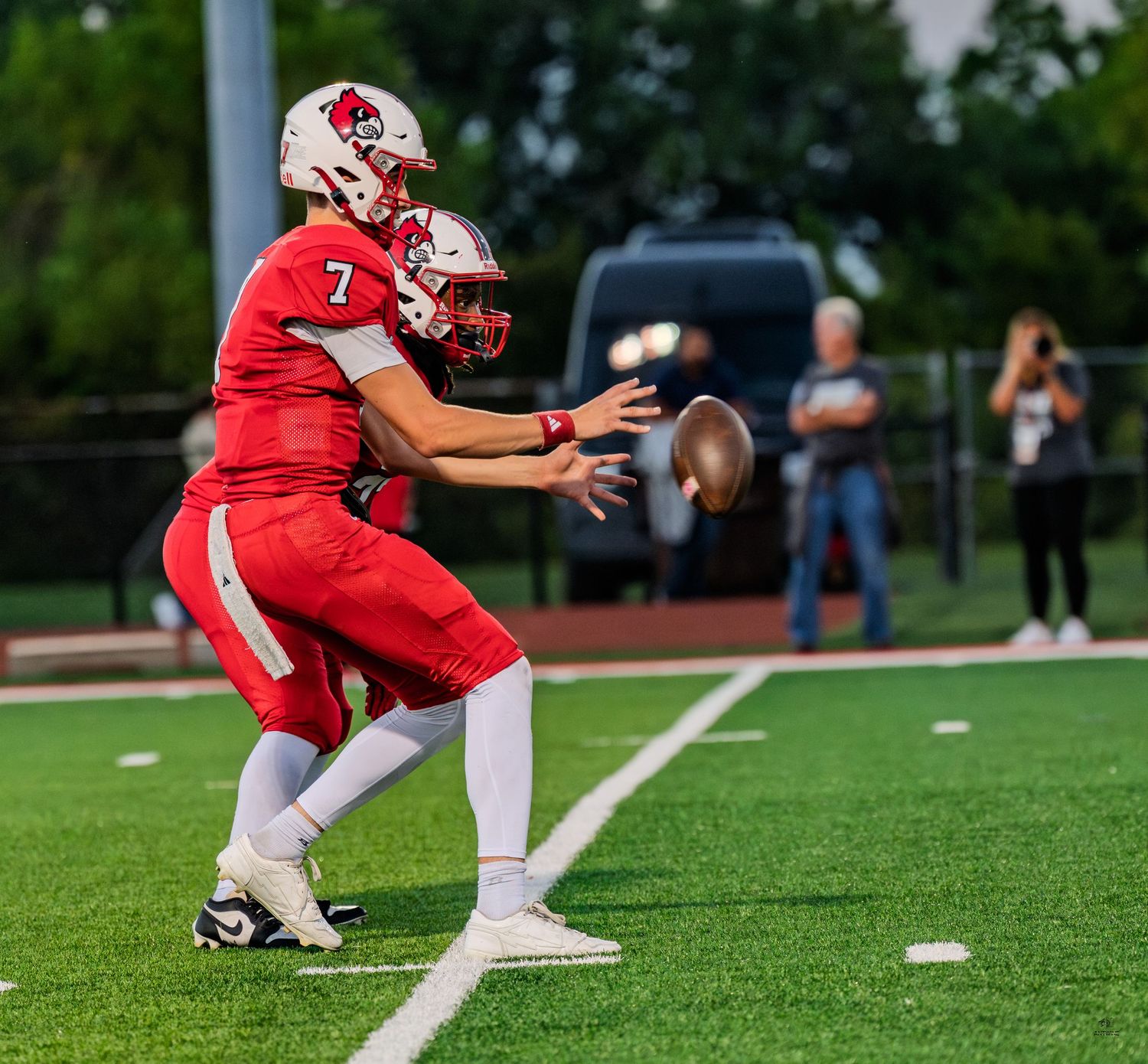 High school football player number 7 in red uniform receives a snap from center during a game.