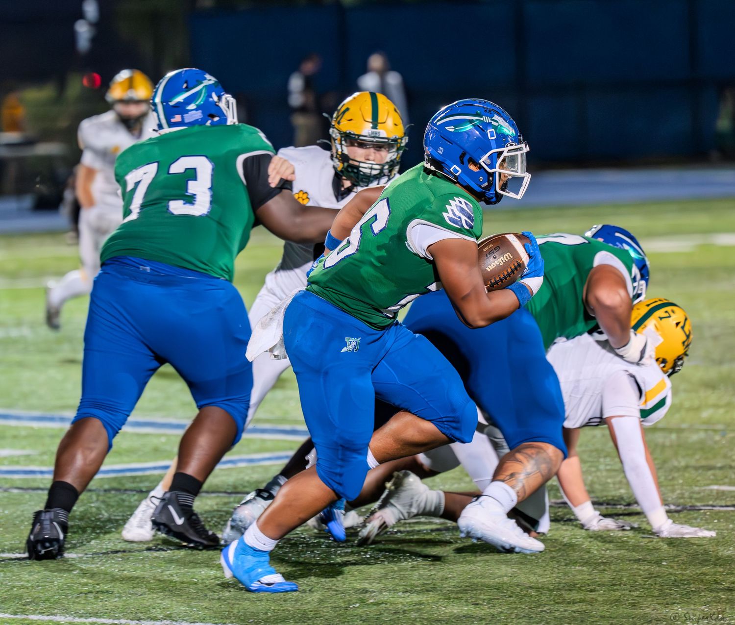 Green and blue Warriors football players execute a running play against yellow-helmeted opponents under stadium lights.