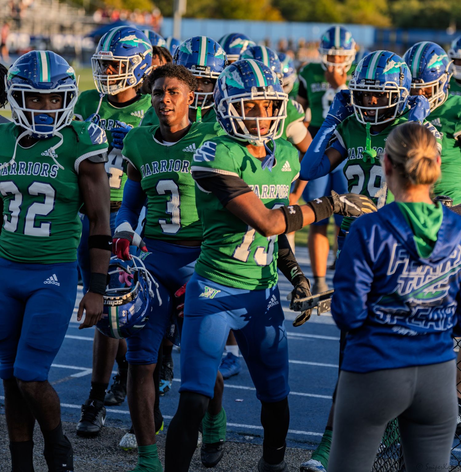 Green and blue uniformed Warriors football players celebrate on the sideline before a game.