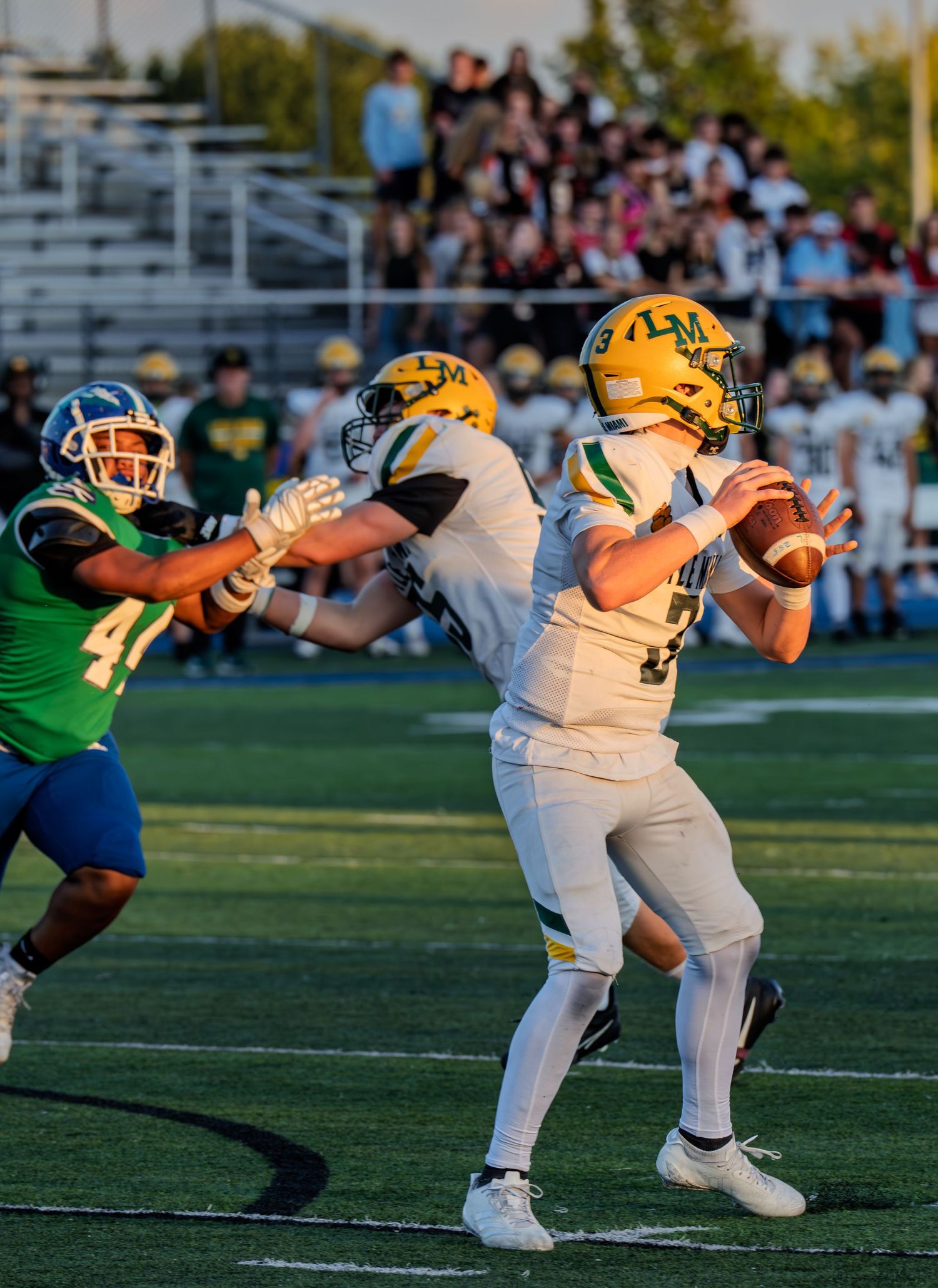 High school quarterback in white and gold uniform prepares to throw while being pressured by a defender.