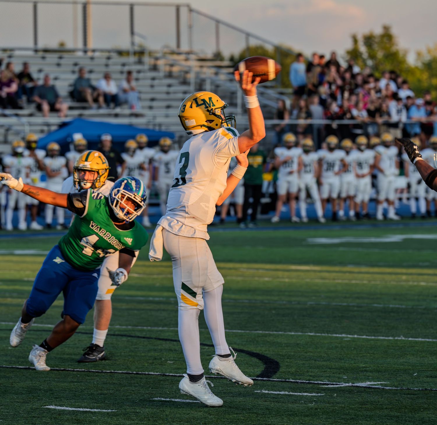 High school football player throws pass while defender rushes in during outdoor evening game.