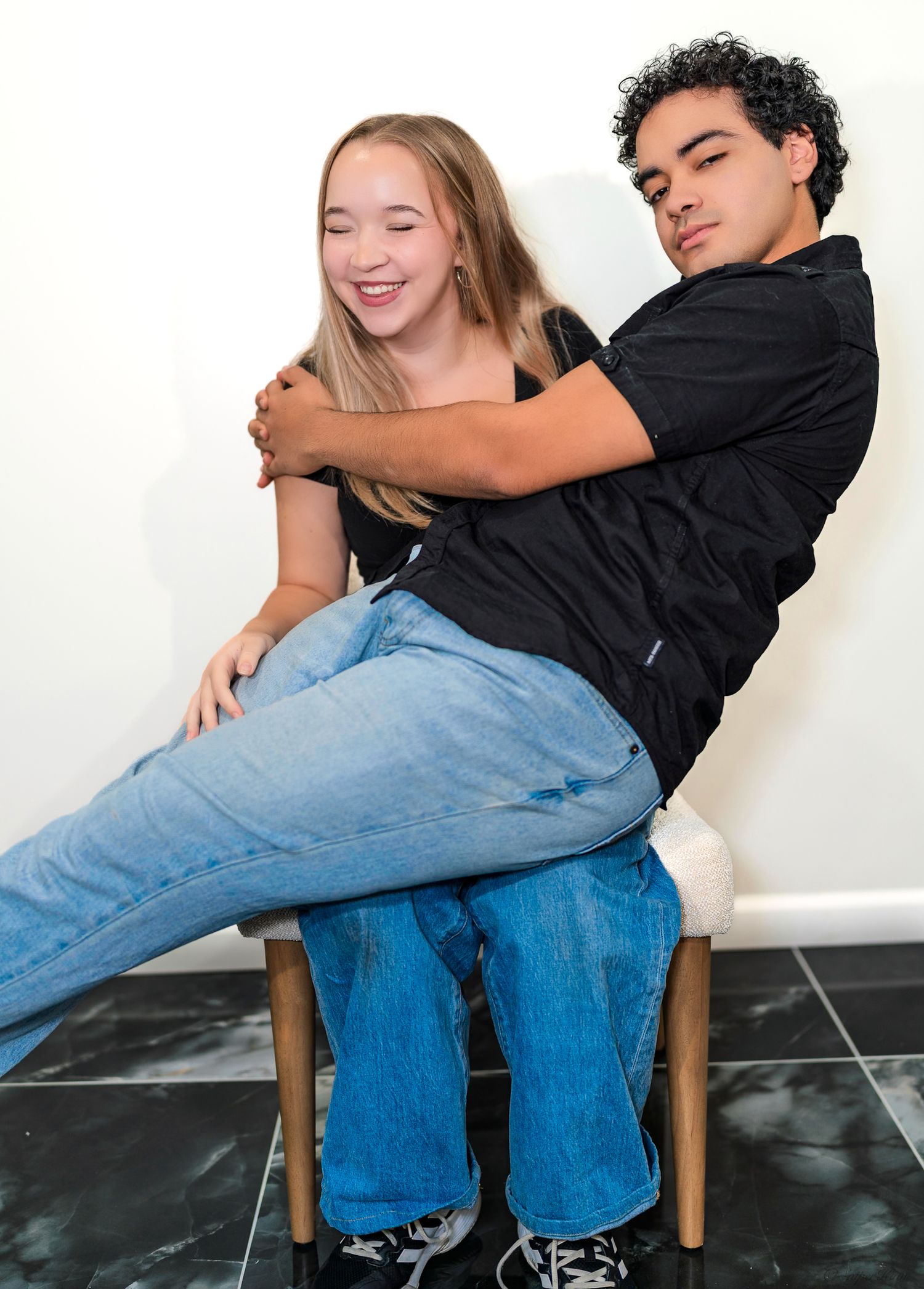 A smiling young woman sits on a bench while a young man leans back against her in a casual portrait.