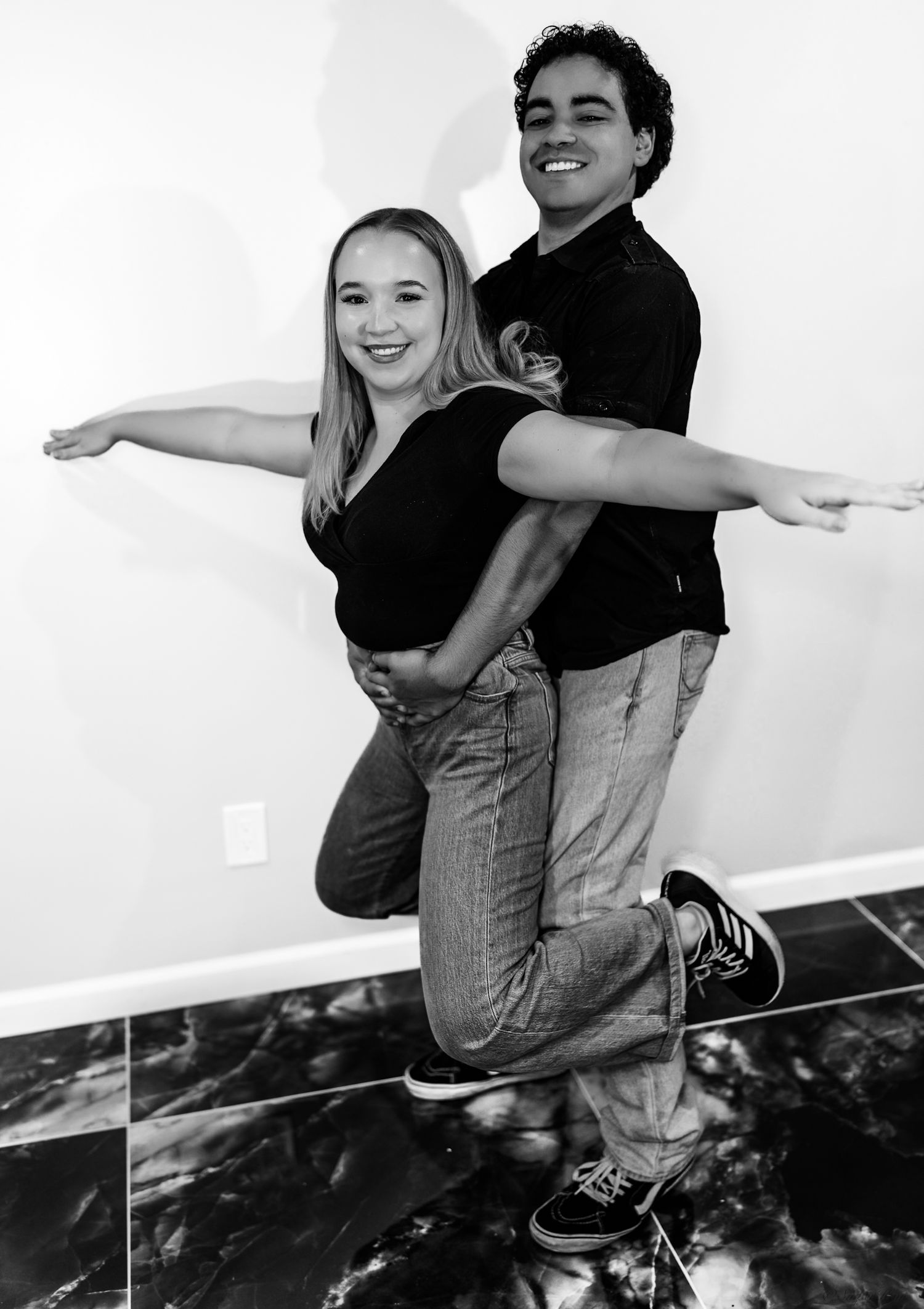 Smiling couple poses playfully with arms outstretched, man lifting woman, in black and white photo.