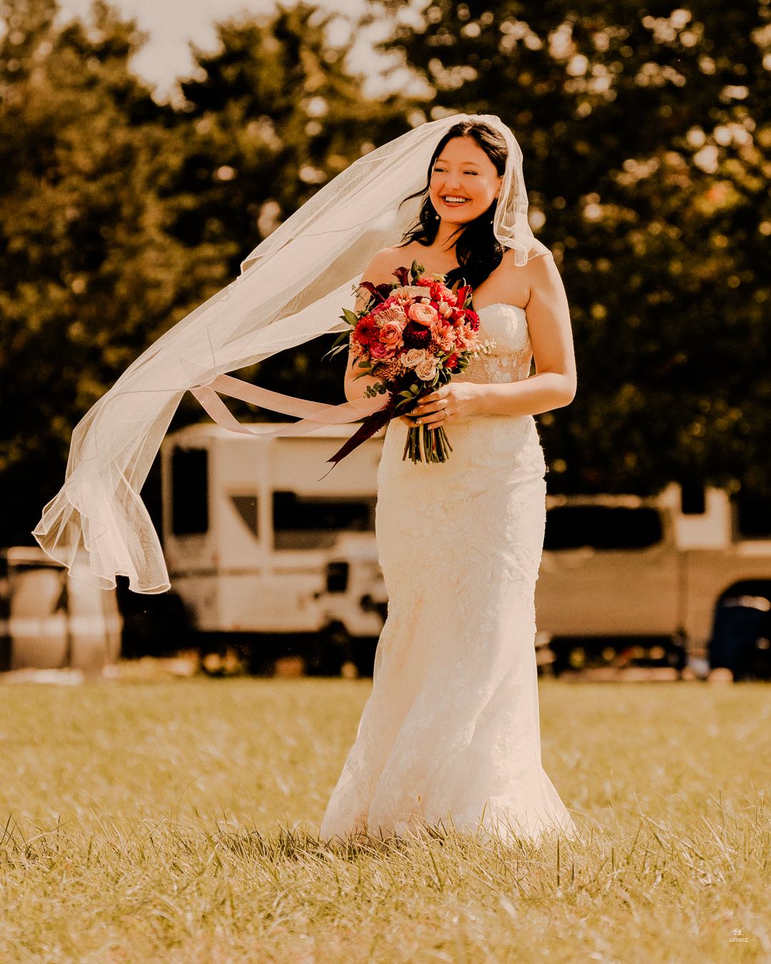 Smiling bride in a white lace gown holds a colorful bouquet as her veil flows in the wind outdoors.