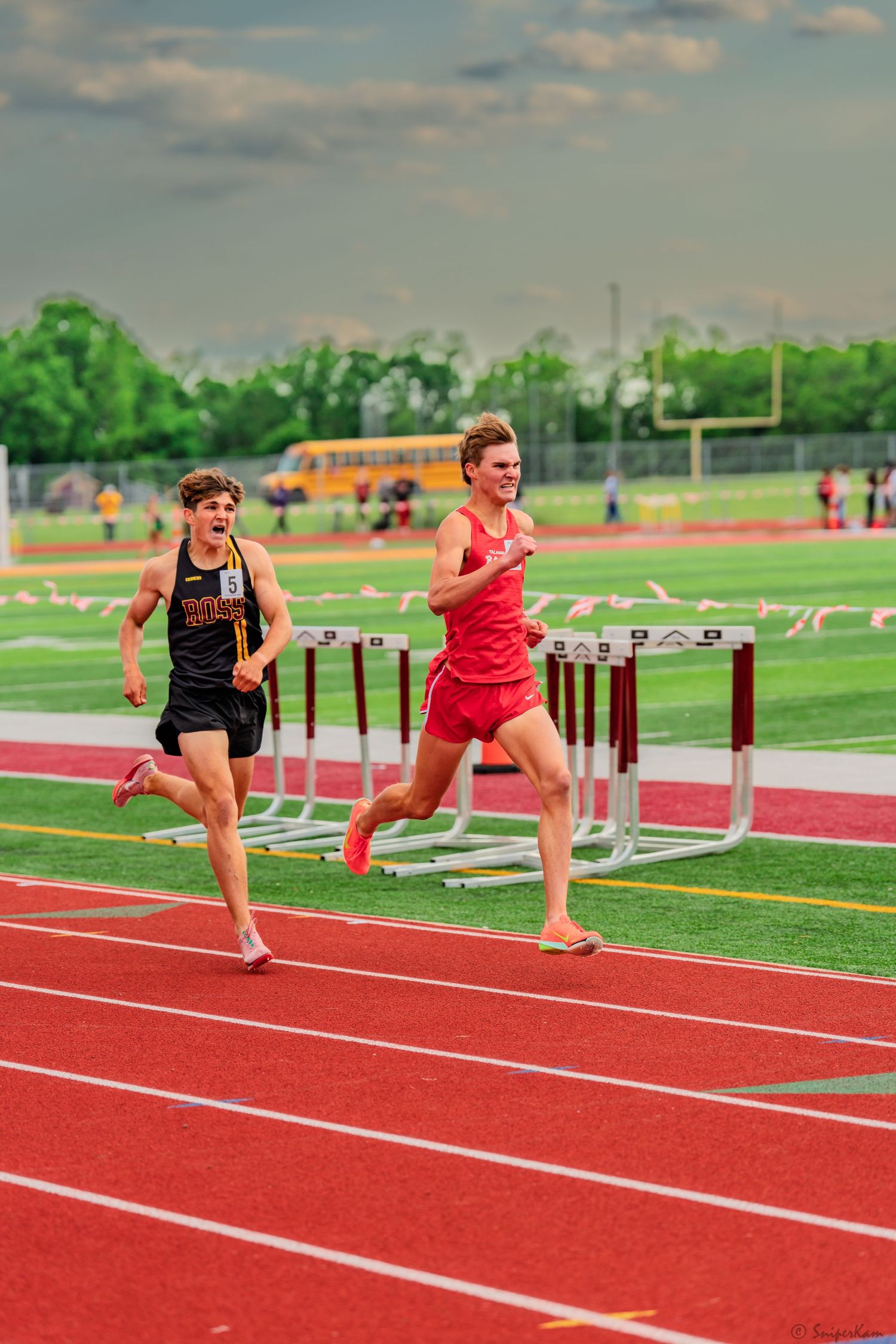 Two male athletes race neck and neck past hurdles on a red outdoor track.