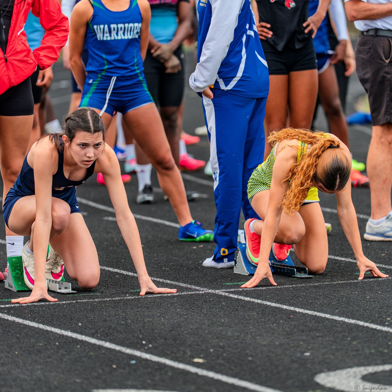 Two female runners in starting blocks prepare for sprint race at high school track meet.