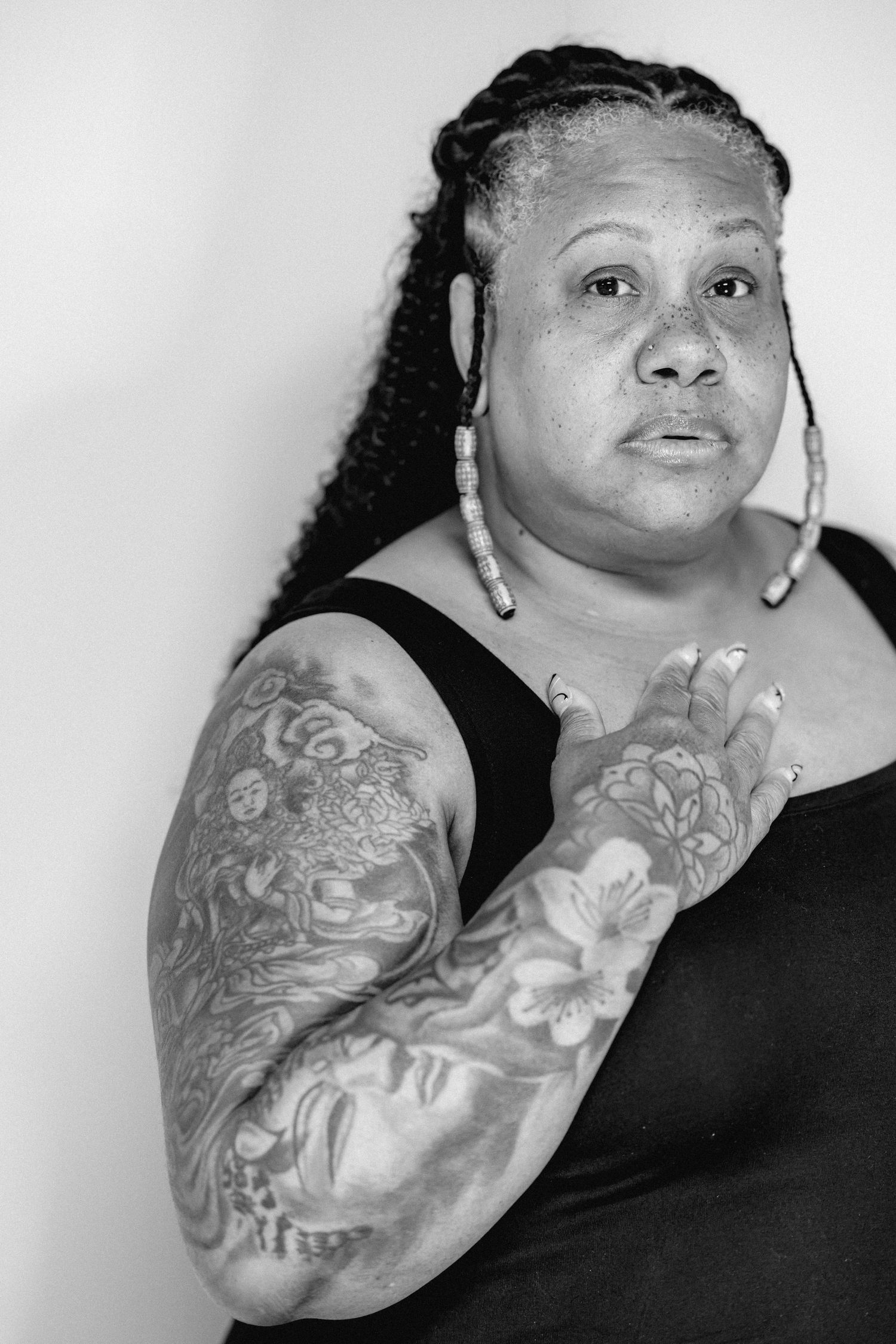 A Black woman with long braids and beaded earrings poses with tattooed arms in a black and white portrait.