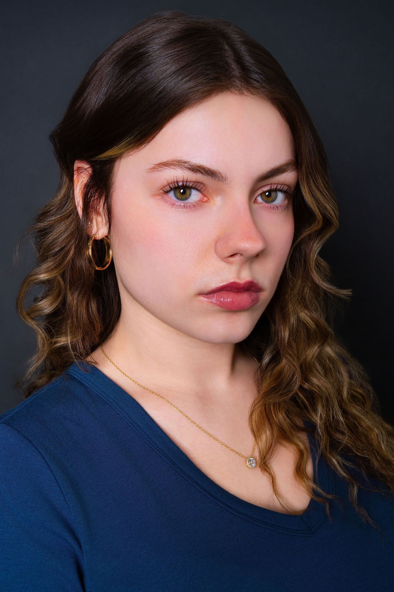 Young woman with curly brown hair, pink eye makeup, and gold necklace in blue top.