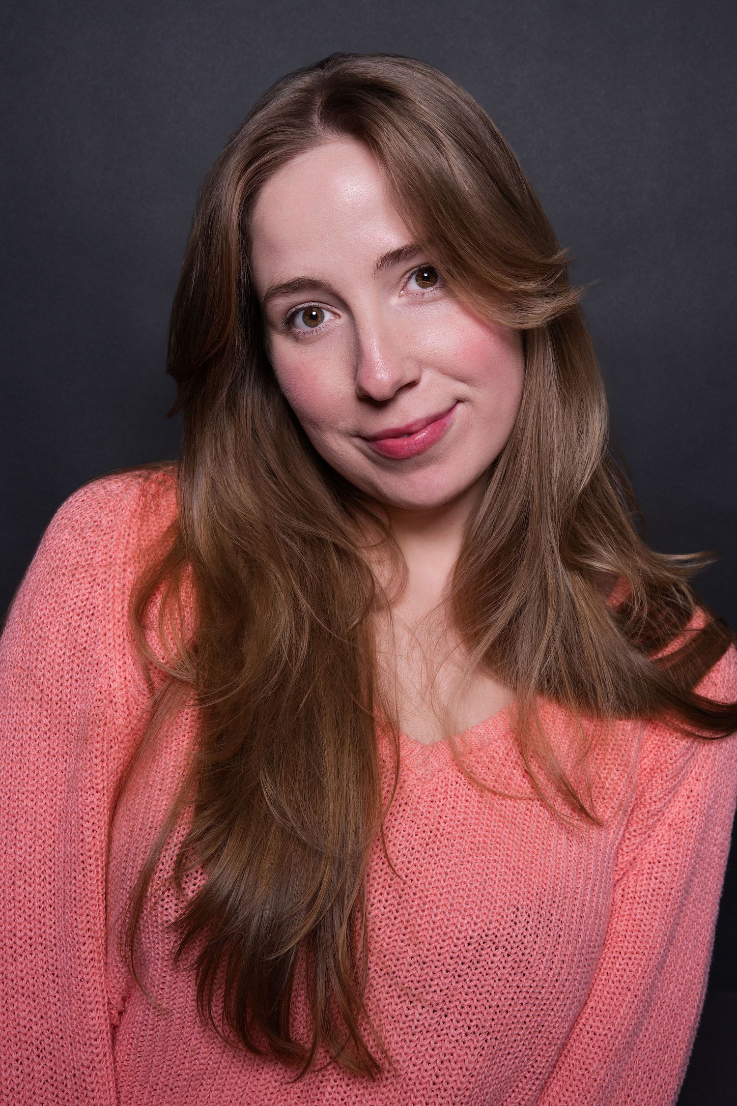 Young woman with long brown hair smiles softly wearing a pink knit sweater in a studio portrait.