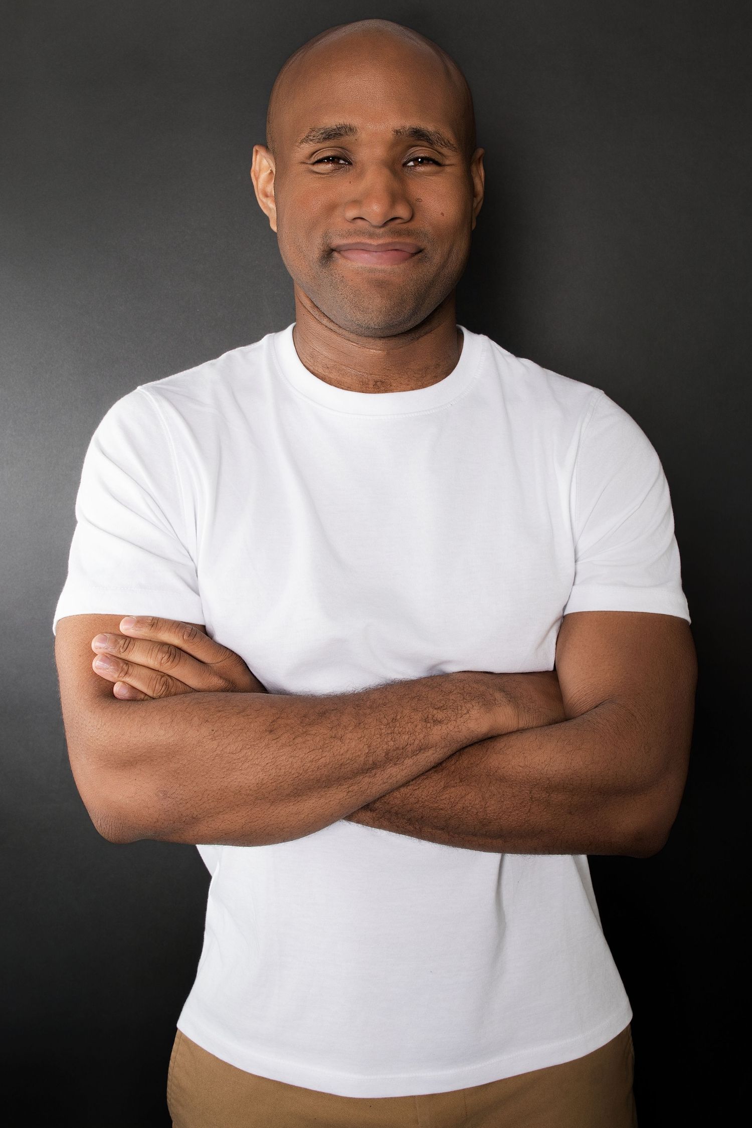 Muscular bald Black man in white t-shirt smiling with arms crossed against dark background.
