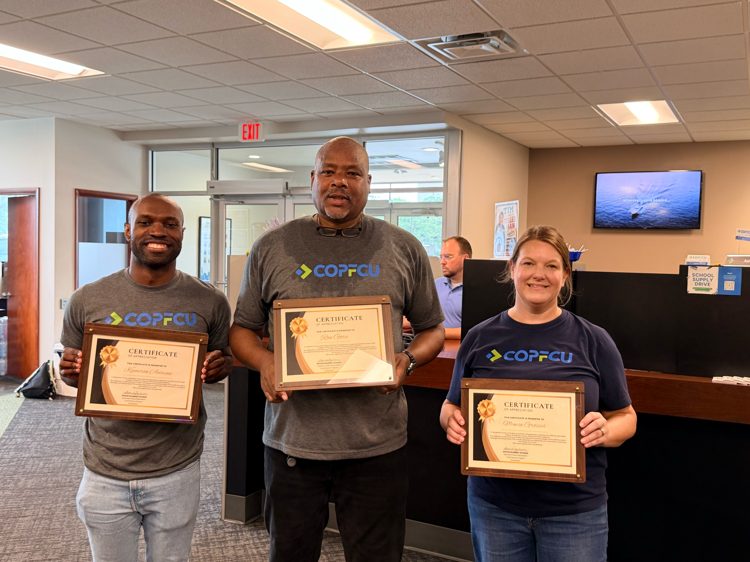 Three COPFCU employees proudly display framed award certificates while wearing branded shirts inside a credit union office.
