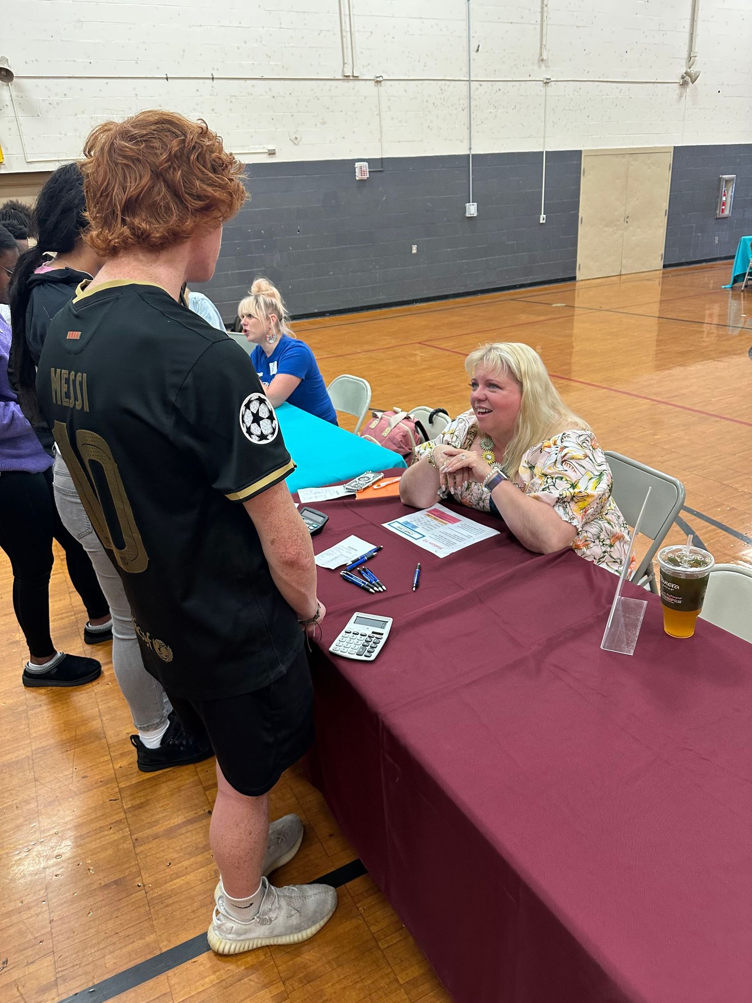 Student in Messi soccer jersey speaks with smiling woman at registration table in school gymnasium.
