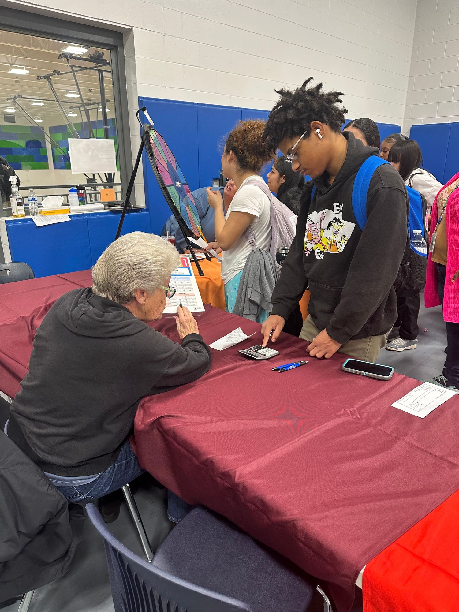 A student uses a smartphone while speaking with a seated adult representative at a maroon-draped career fair table.
