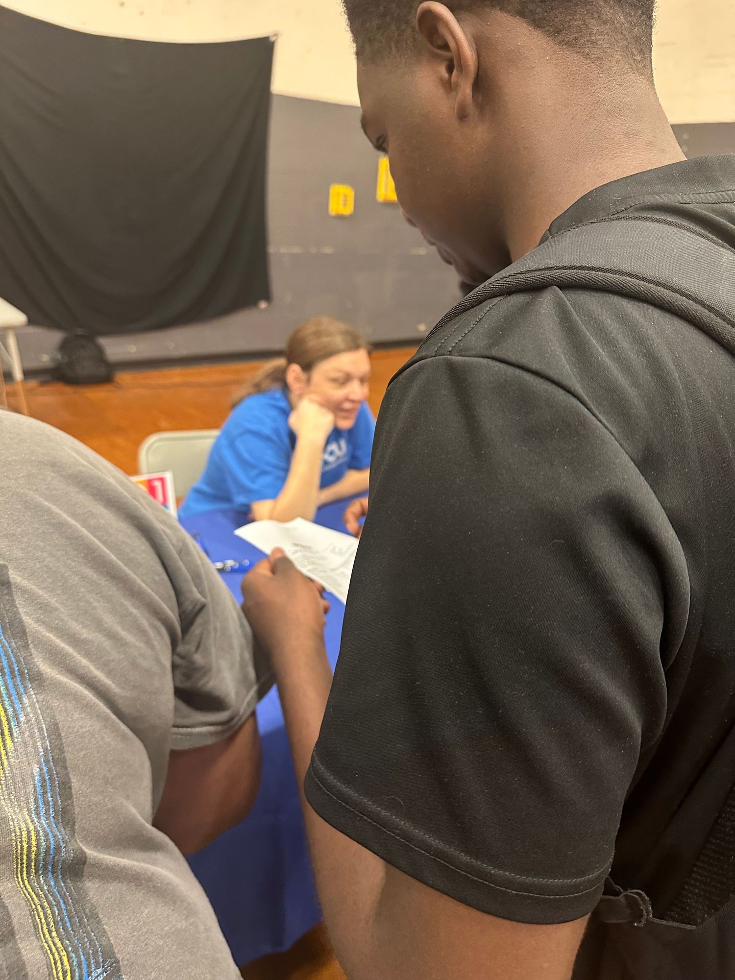 Students review documents at a school career fair table with a female advisor in a blue shirt seated nearby.