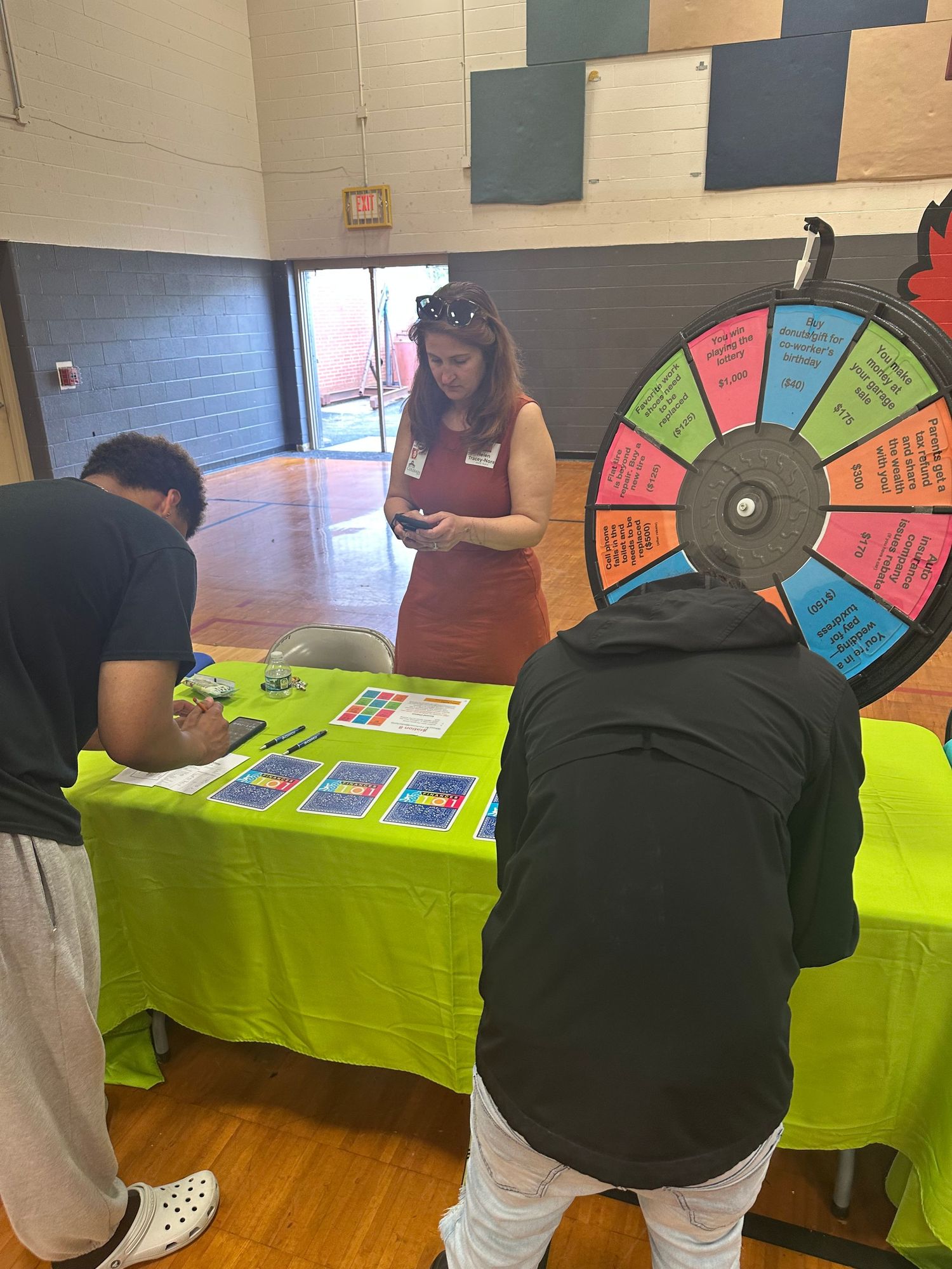 Students spin a colorful prize wheel at a green table booth during a school event in a gymnasium.