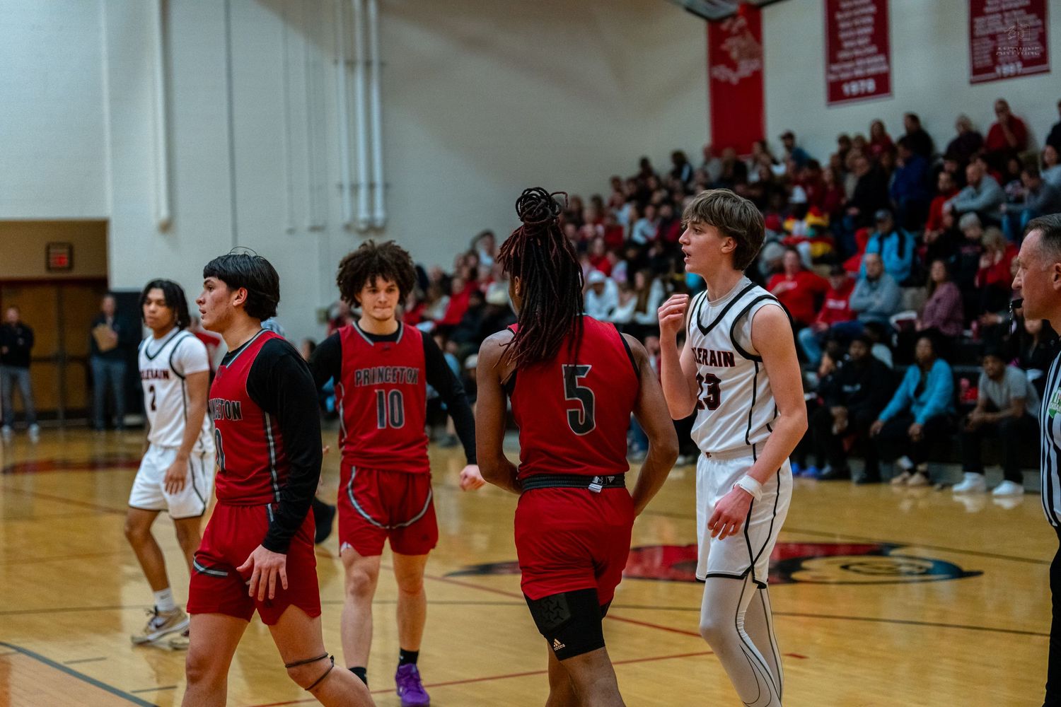 Princeton basketball team players in red jerseys walk on court during high school game with packed stands.