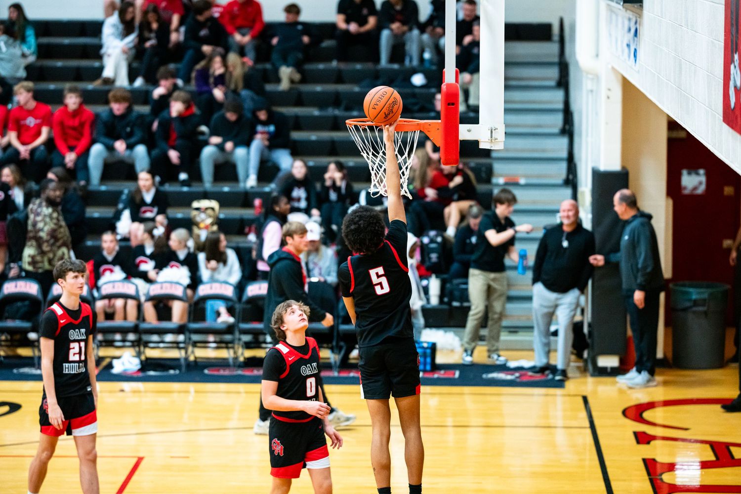 High school basketball player number 5 attempts a layup during a game with fans watching in the stands.