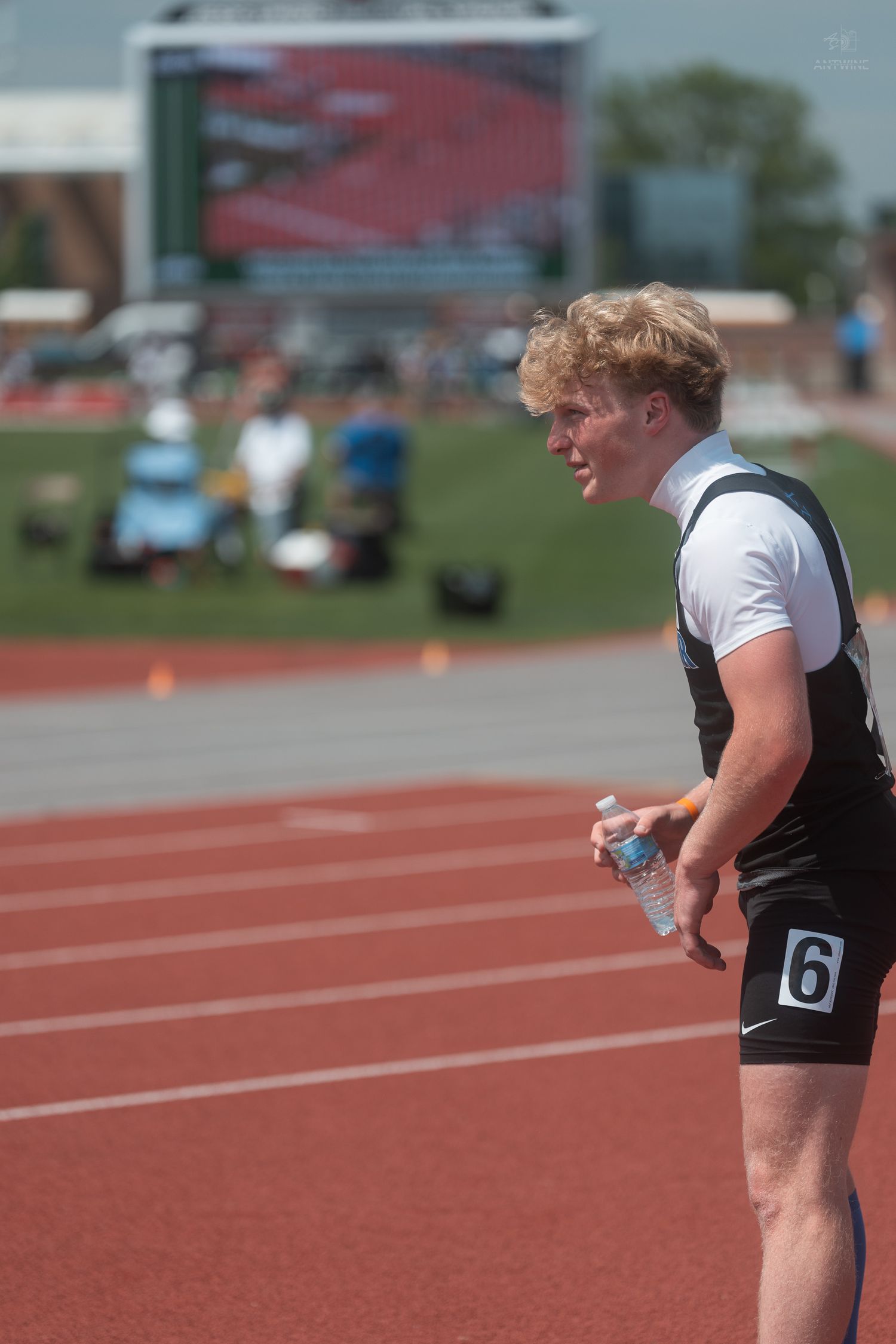 Blonde male athlete in black uniform holds water bottle on red track at outdoor meet.