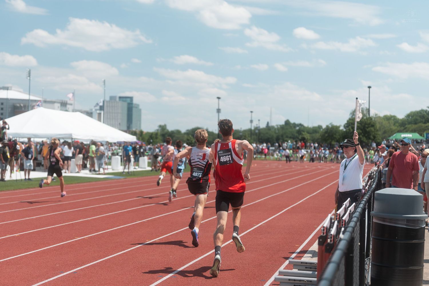 Runners competing in an outdoor track race on a red track with spectators lining the sidelines on a sunny day.