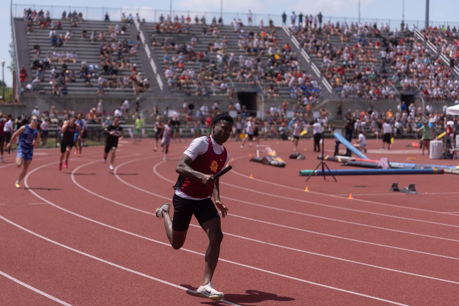 A runner in a red jersey competes on a track at a large outdoor track and field championship.