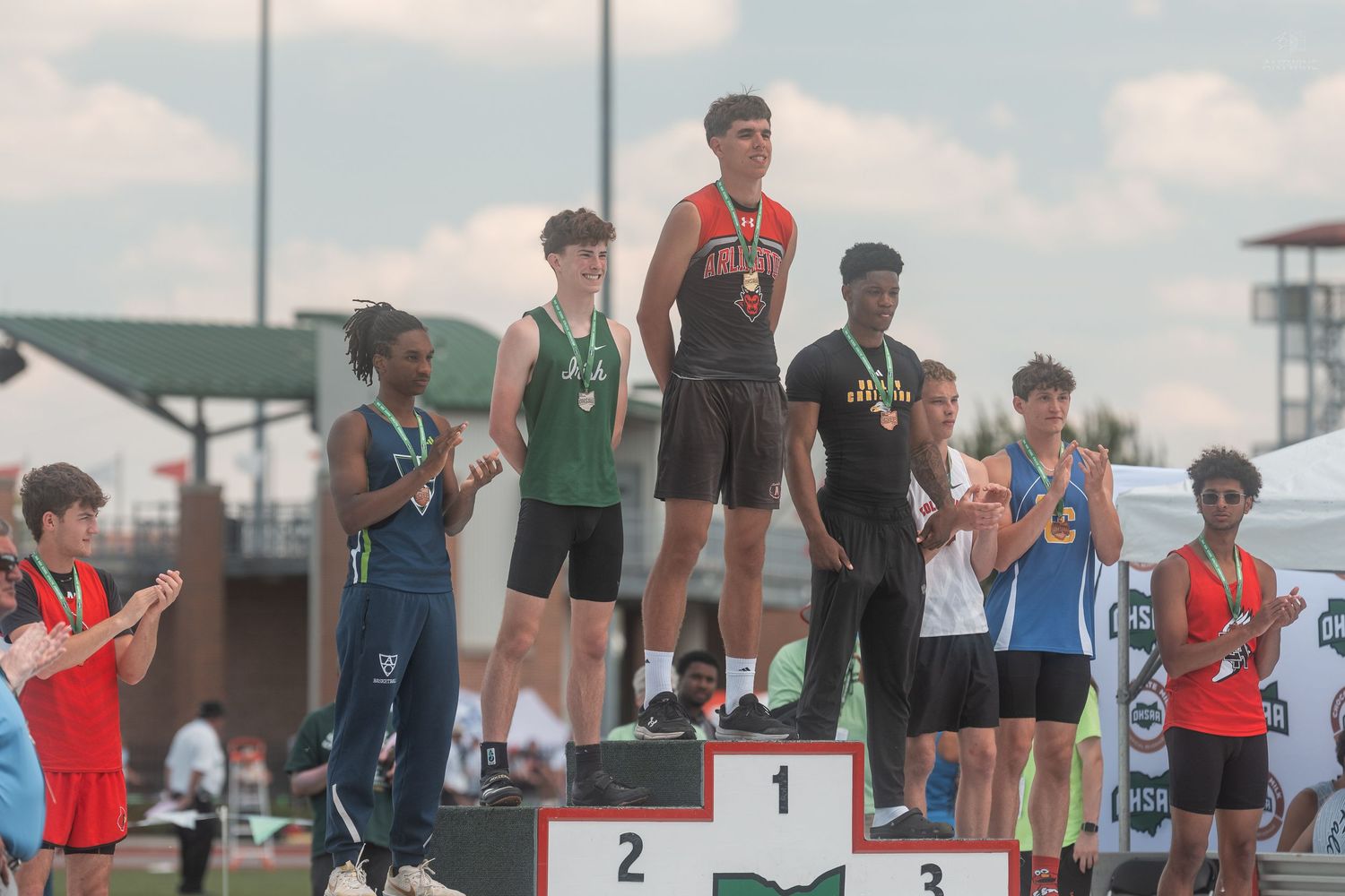 Young athletes celebrate on a numbered podium at an outdoor track and field championship meet on a sunny day.
