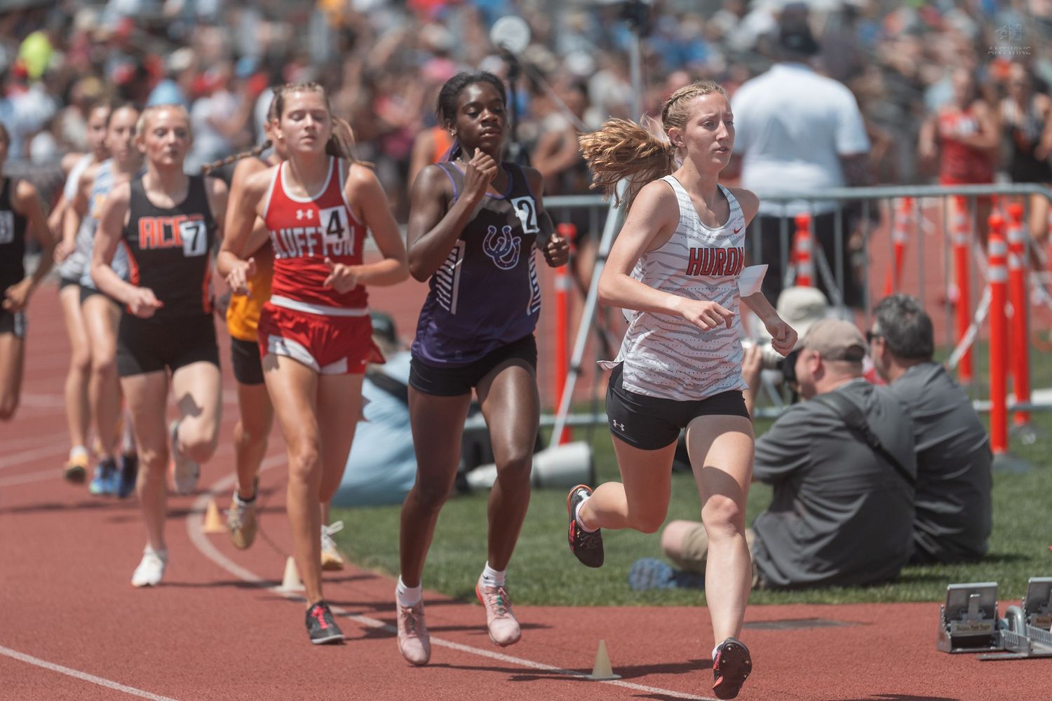 Young female runners compete in a middle distance track race at a sunny outdoor high school track meet.