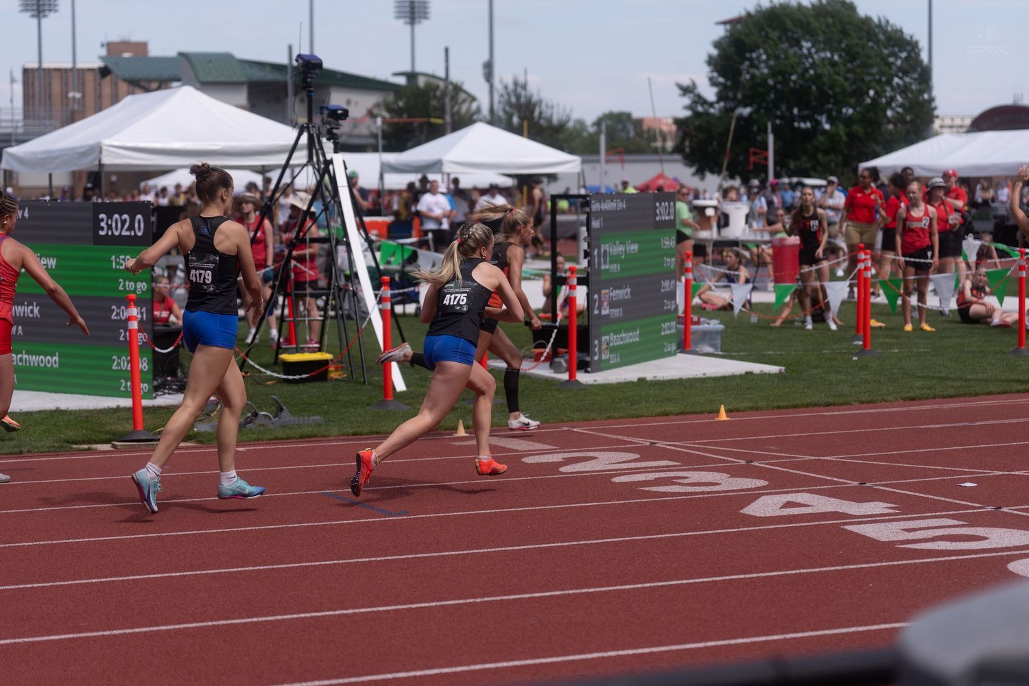 Female athletes sprint toward the finish line at an outdoor track and field competition on a red track.