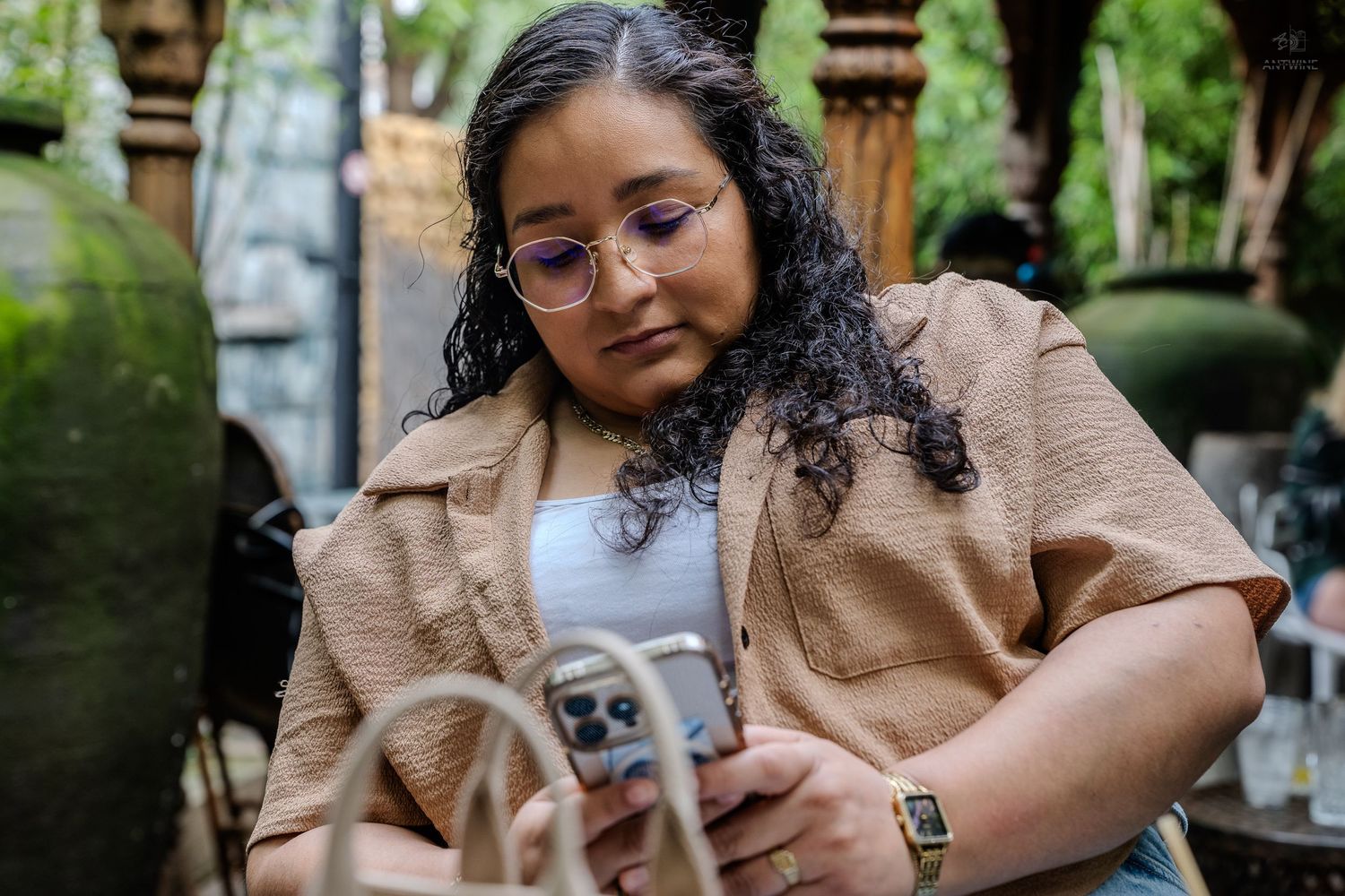 Woman with curly hair and glasses using smartphone outdoors at garden cafe.