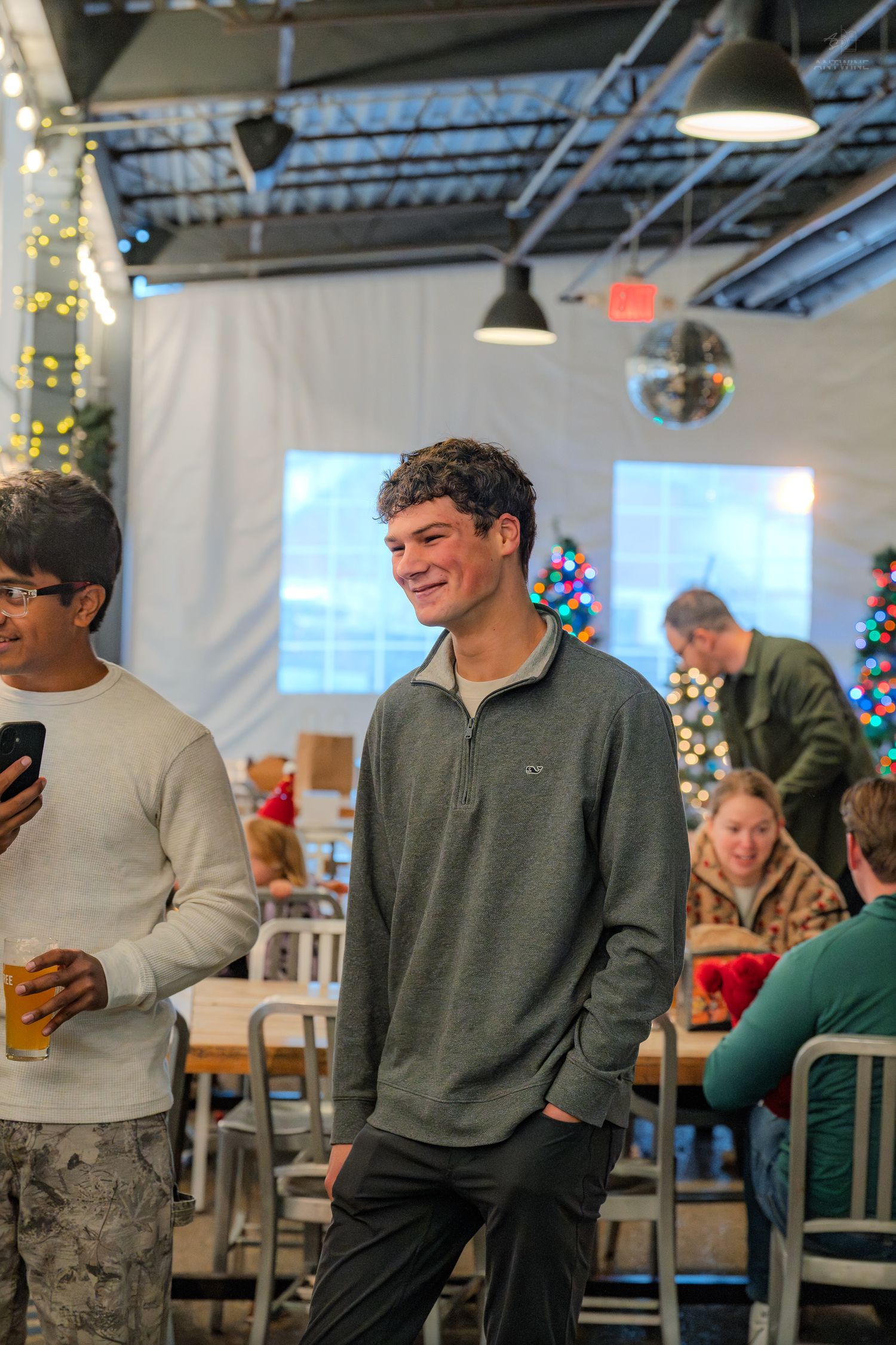 A smiling young man in a gray quarter-zip stands at a holiday party with a Christmas tree and string lights nearby.