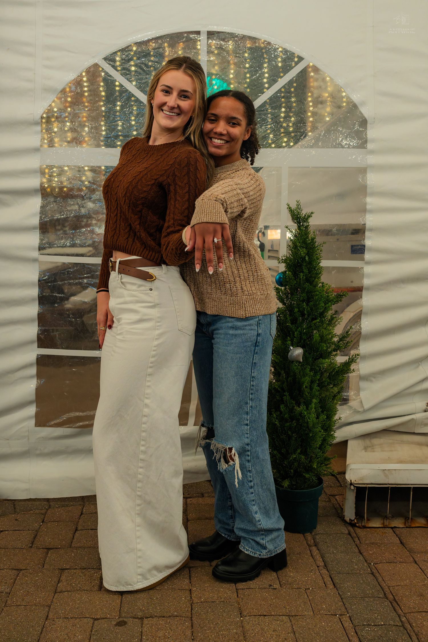 Two young women posing back-to-back inside a white tent decorated with fairy lights and a small tree.