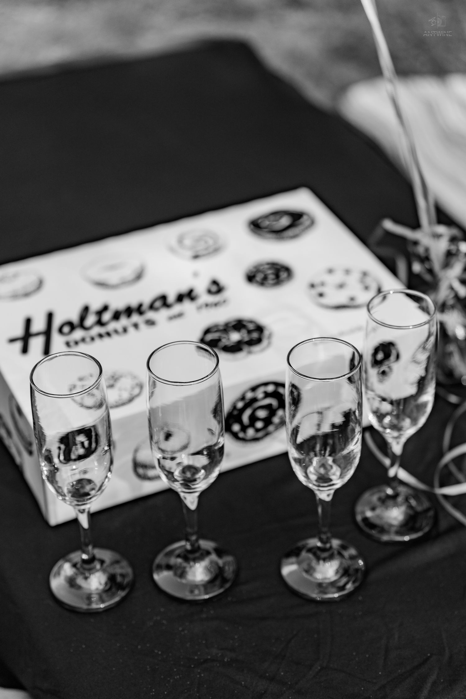 Black and white photo of champagne flutes lined up beside a Holtman's Donuts box on a dark tablecloth.
