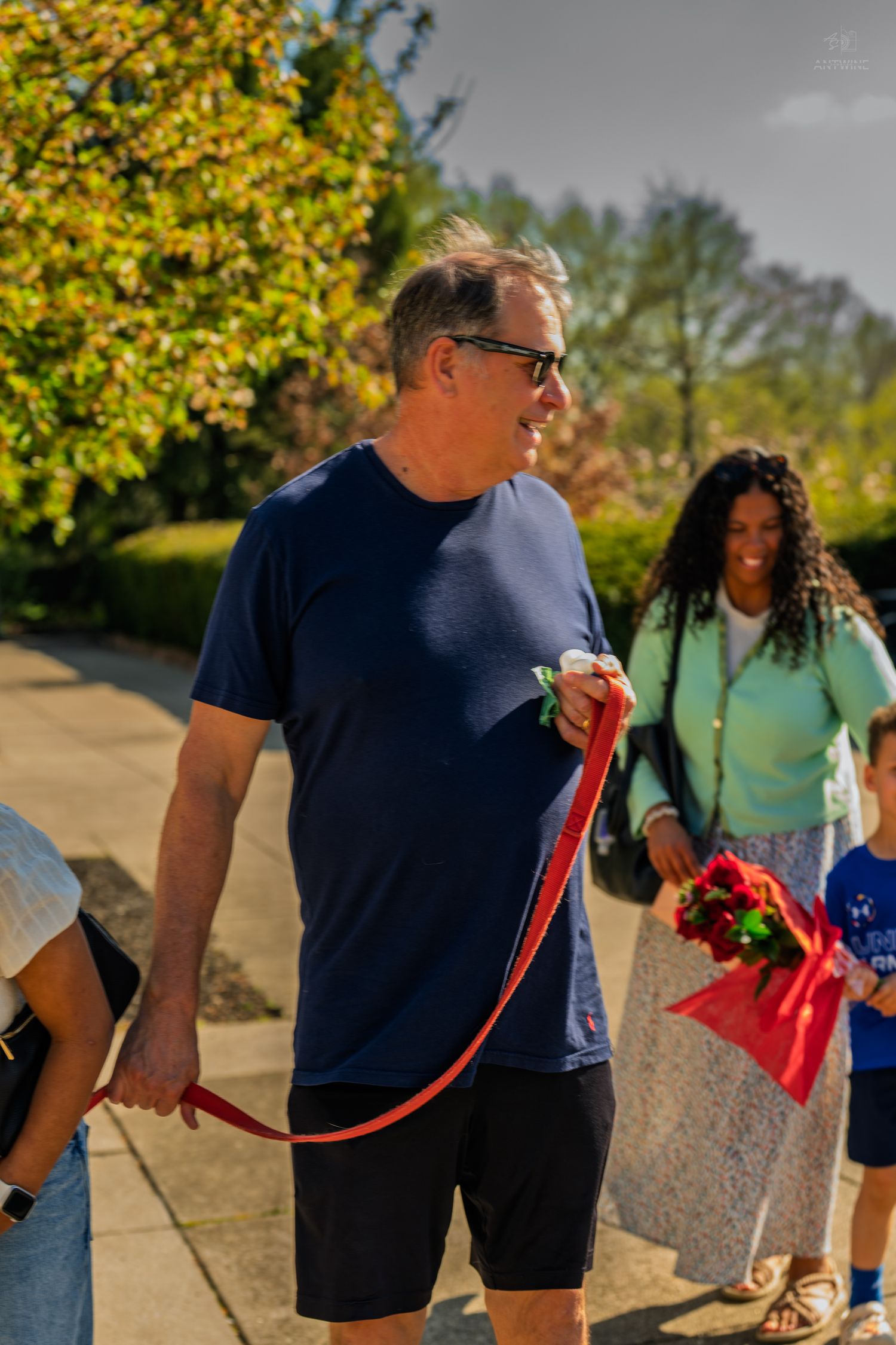 Smiling man in navy shirt walks with red dog leash outdoors, woman and child with red roses behind him.