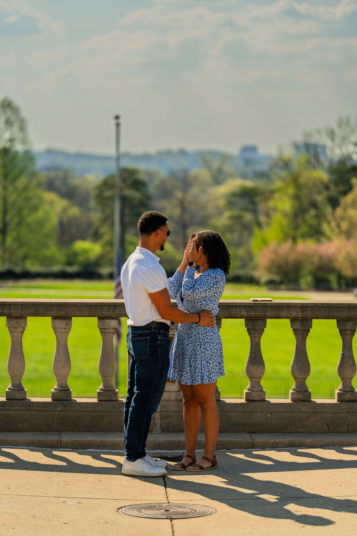 Romantic couple embracing by classical stone balustrade with lush green park landscape behind them.