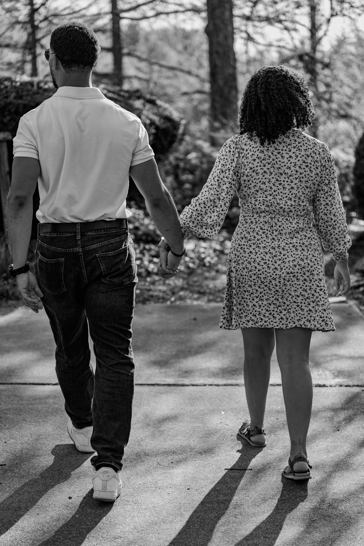 Black and white rear view of a couple holding hands strolling along a sunlit park pathway.