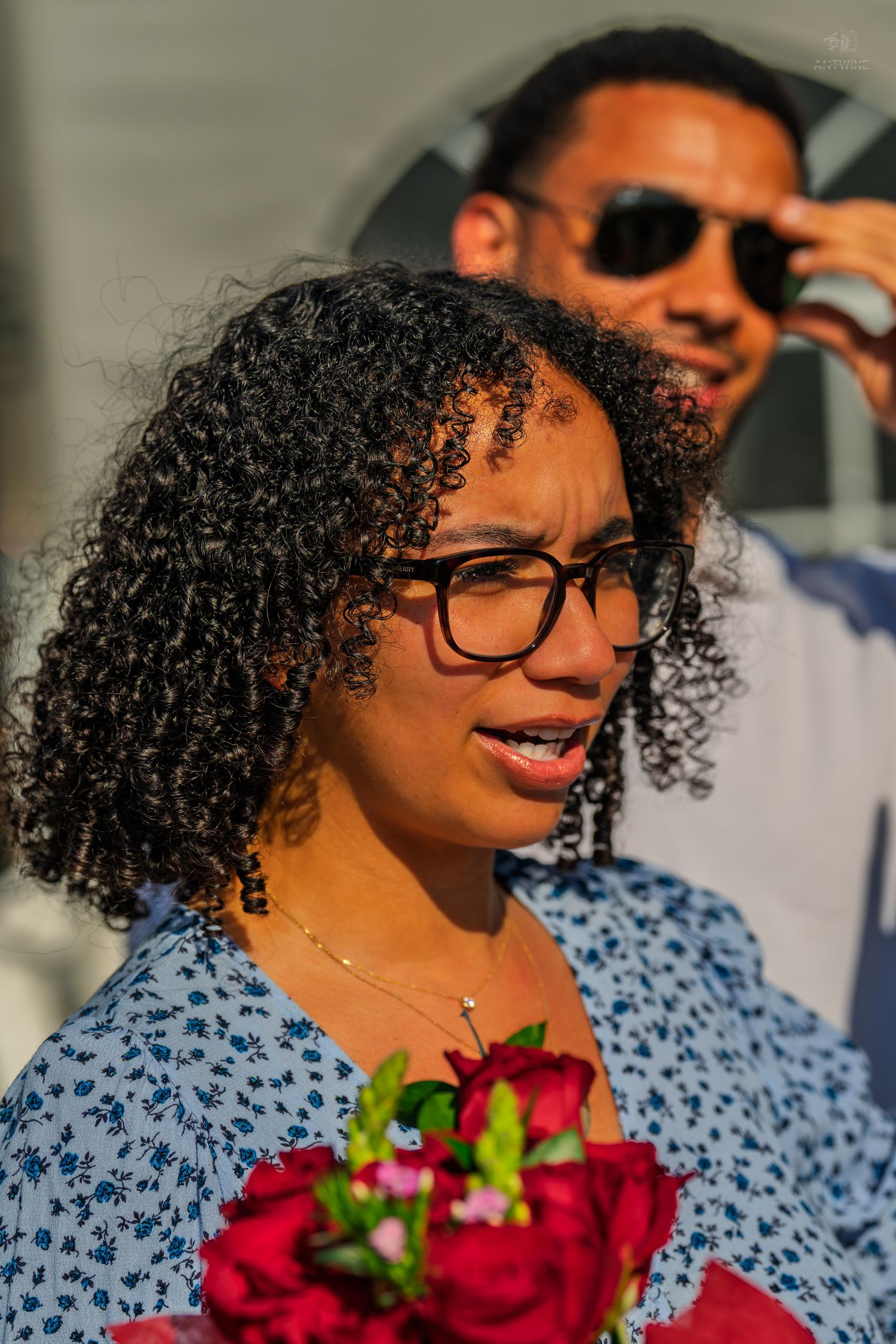 A woman with curly hair and glasses holds red roses bouquet outdoors, man in sunglasses visible behind her.