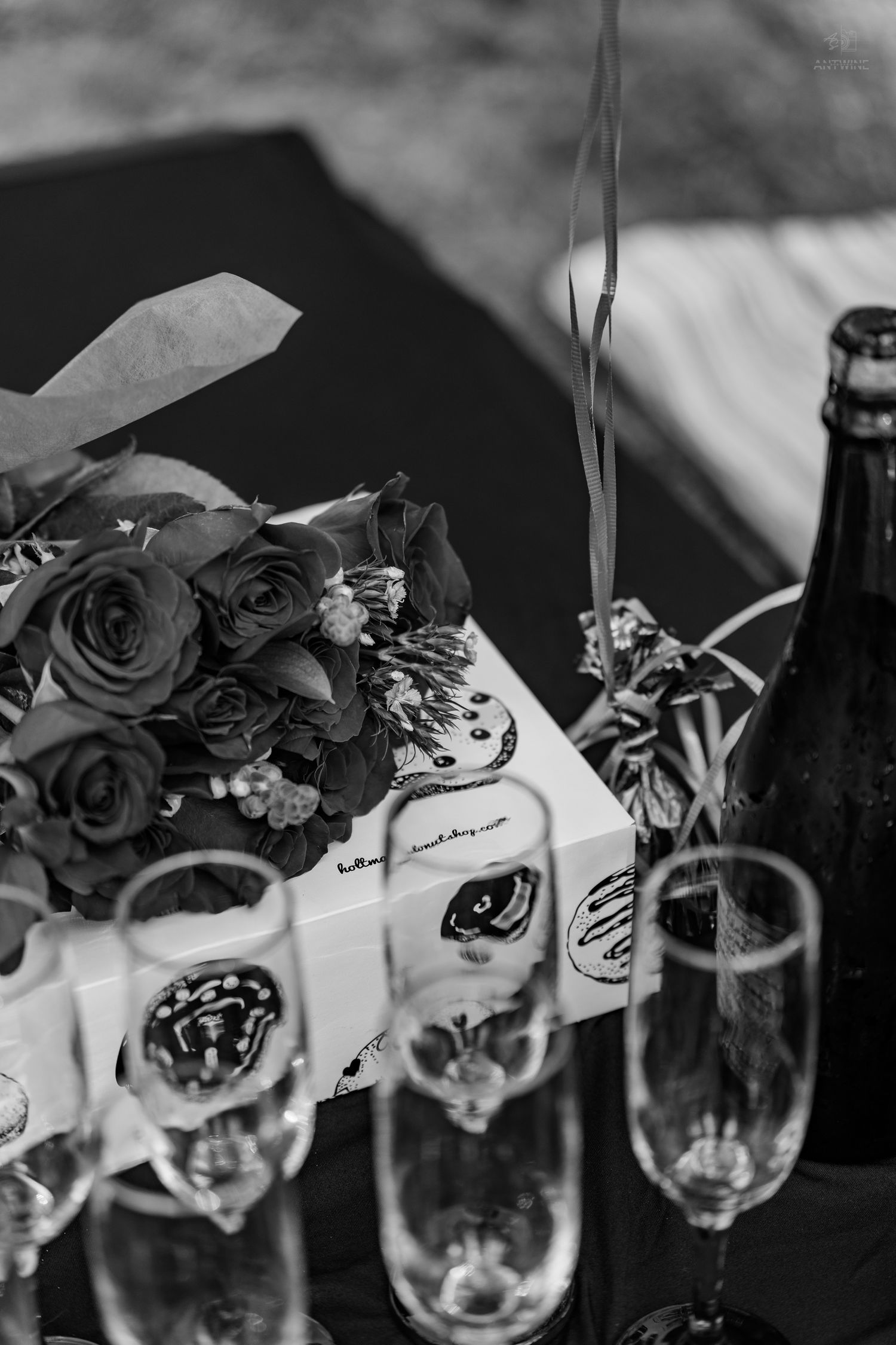 Black and white image of rose bouquet, champagne flutes, and bottle on a decorated table.