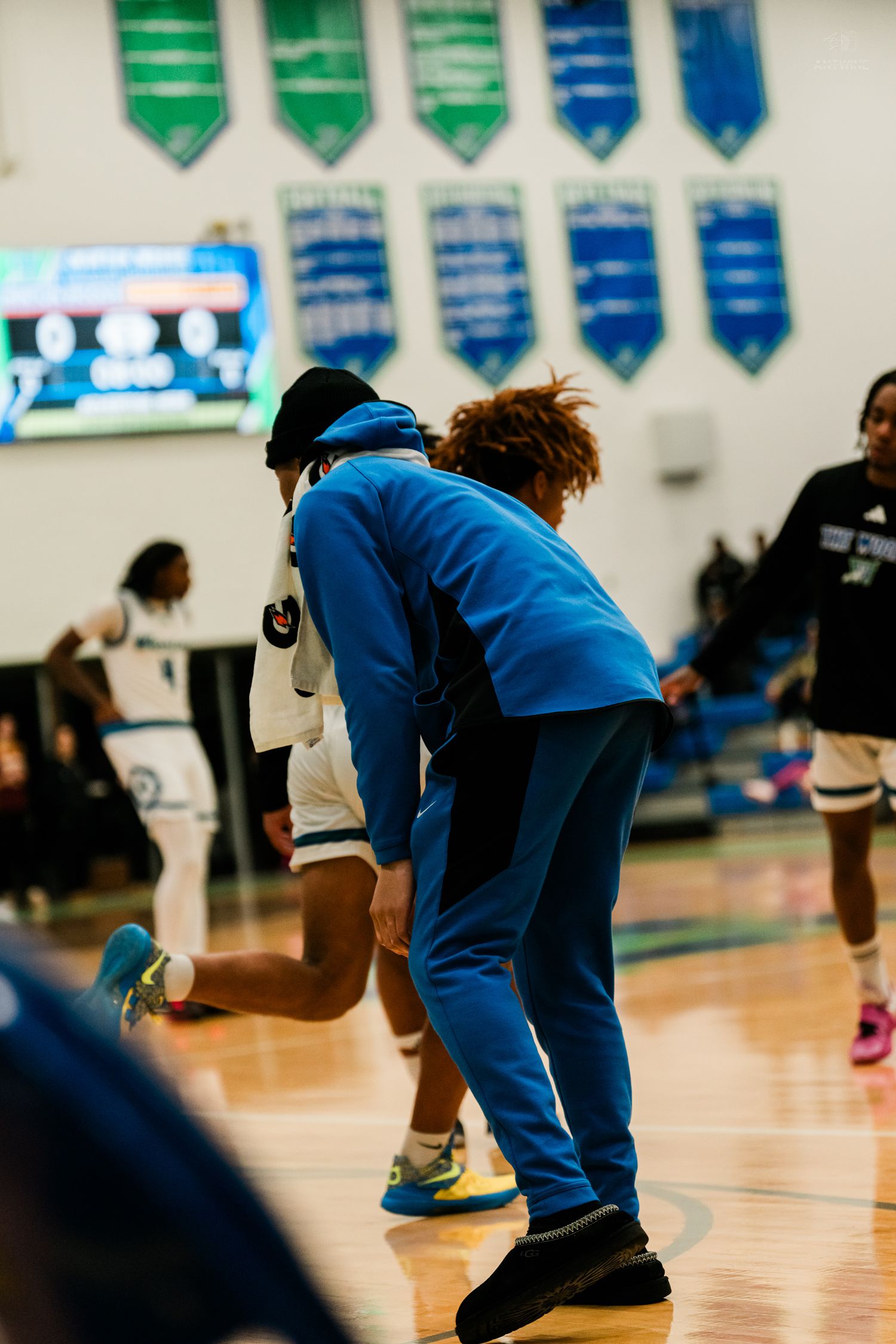 Basketball coach in blue hoodie bends down to speak with player during pregame warmups in gym.