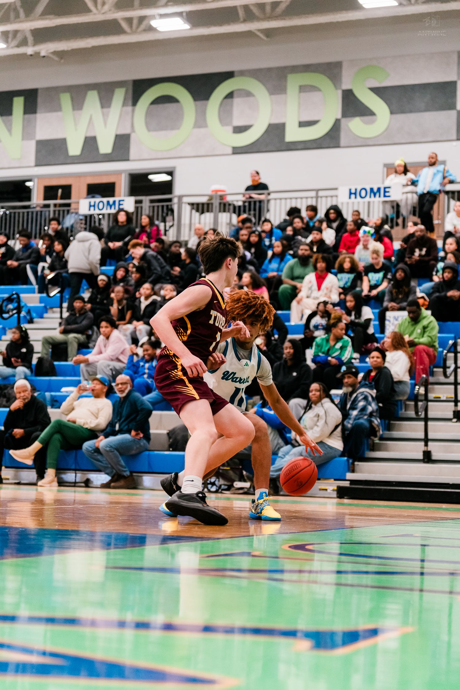 Warriors player drives baseline against maroon-uniformed defender in packed gymnasium.
