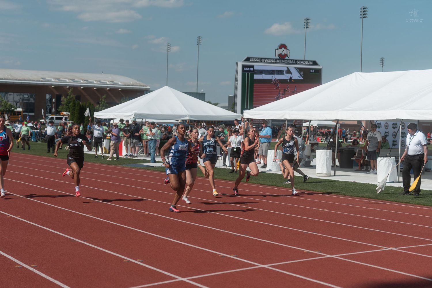 Female athletes sprint toward the finish line on a red track at an outdoor high school track competition.