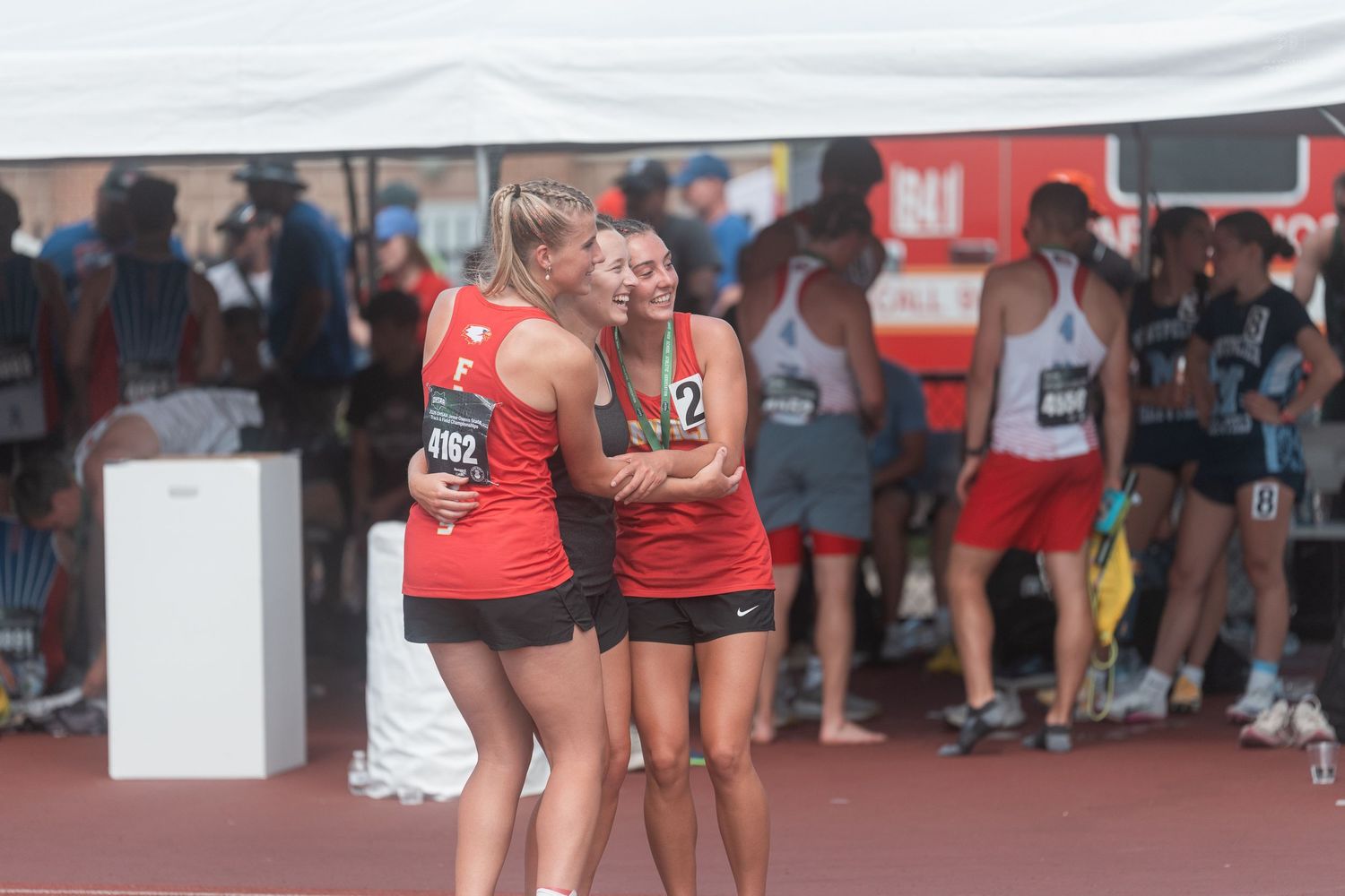 Female track athletes embrace under a white tent after competing in a track and field event.