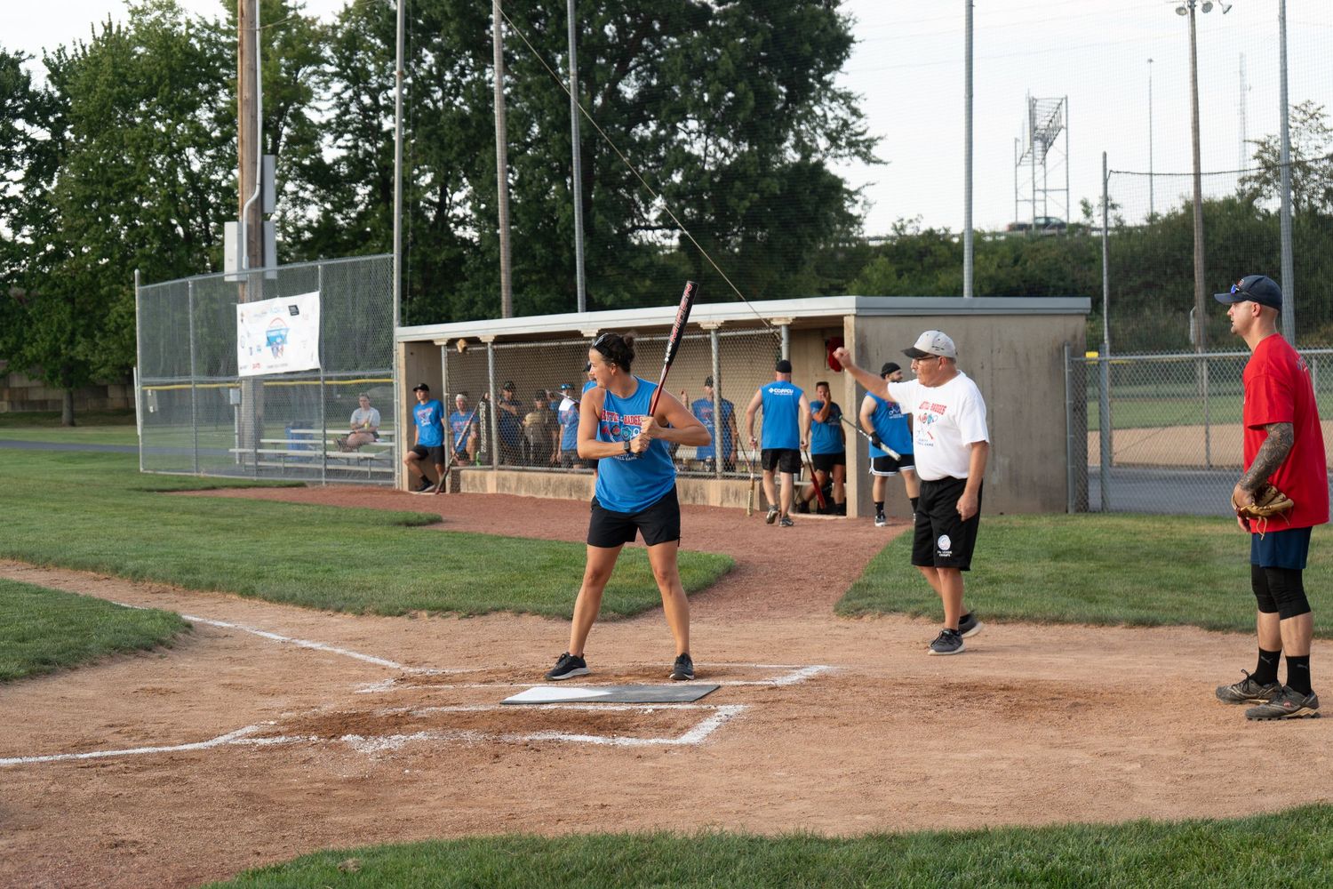 Batter in blue shirt preparing to swing at home plate during an outdoor recreational baseball game at dusk.